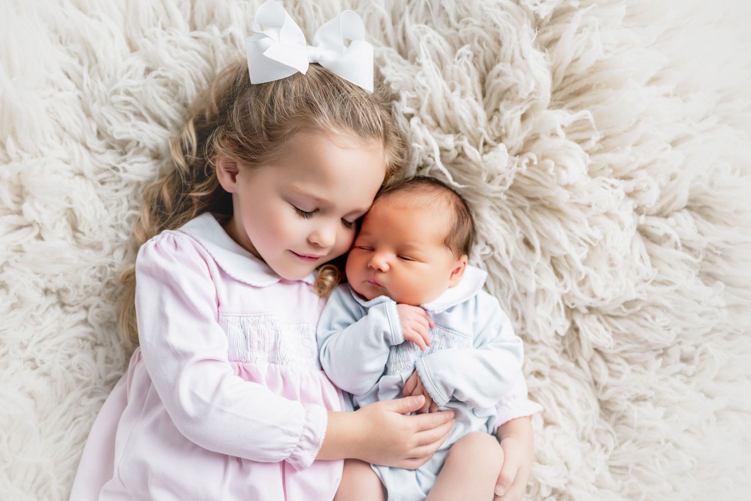 Young girl in pink dress tenderly holding newborn baby sibling on fluffy white rug.