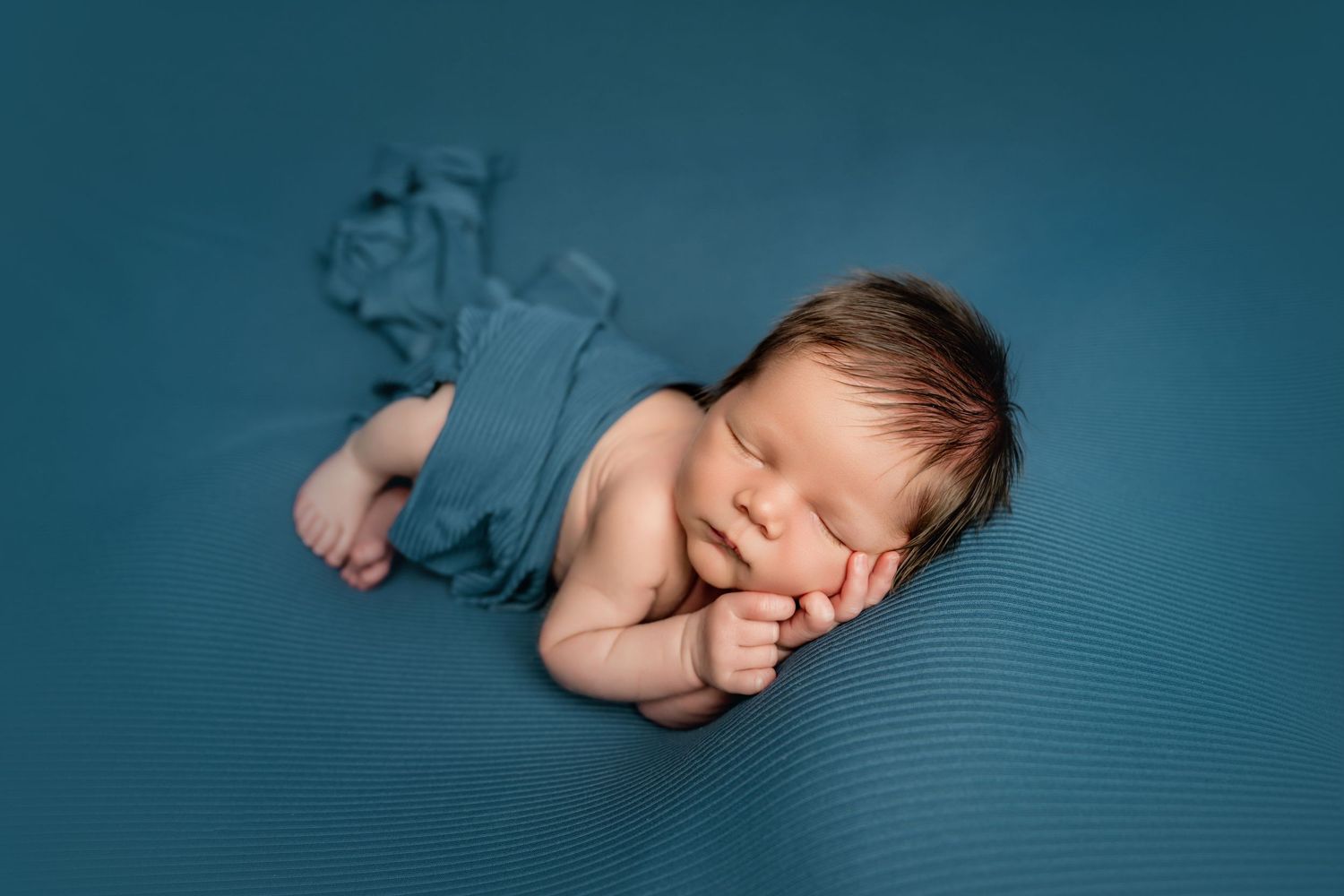 Peaceful newborn baby sleeping wrapped in teal fabric on a matching teal textured background.