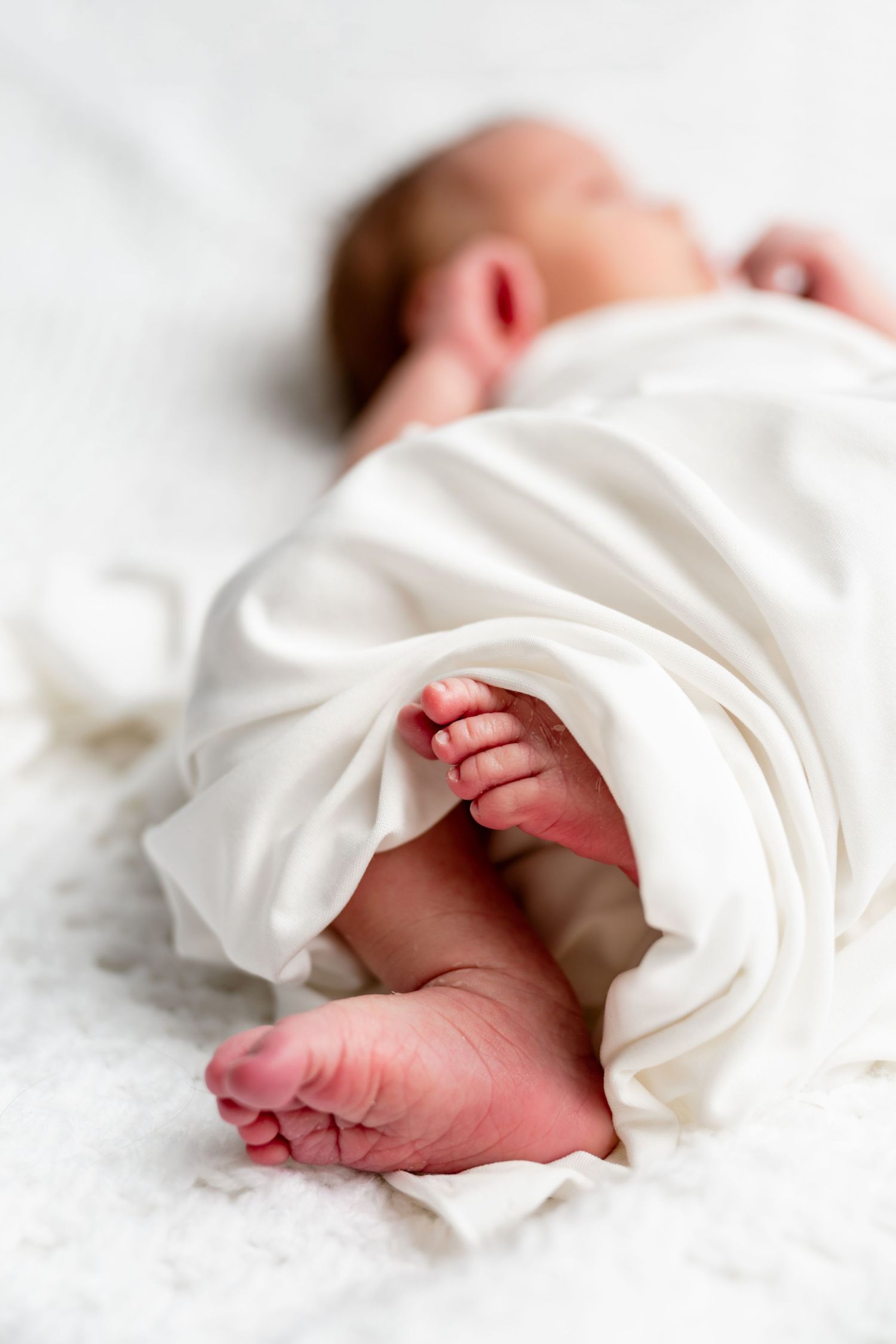 Close-up of a sleeping newborn baby wrapped in a soft white blanket with tiny pink feet visible.