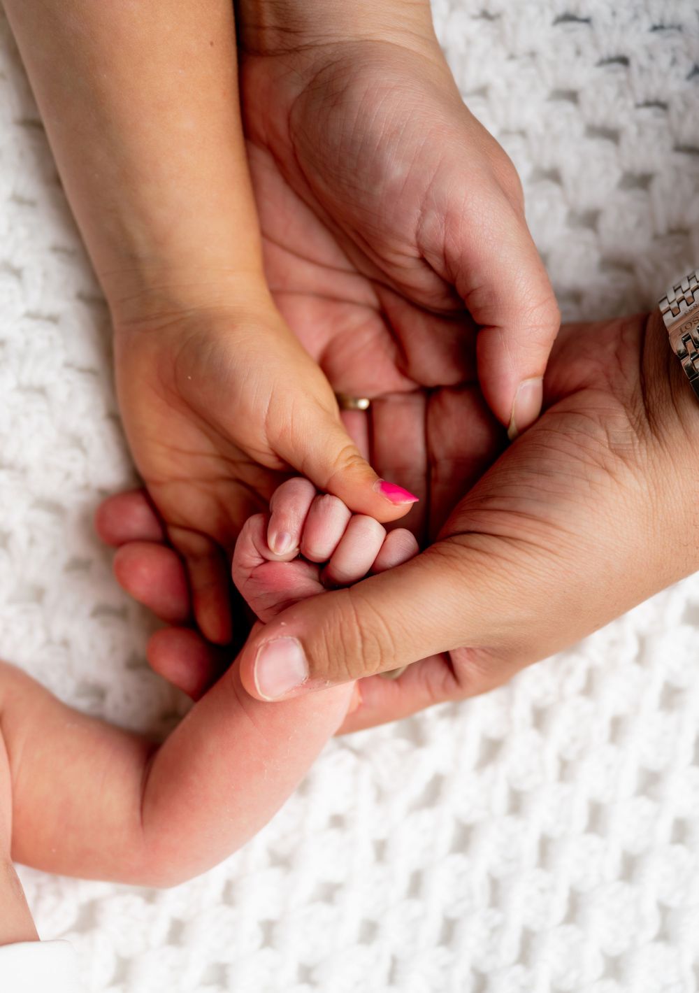 A newborn baby's tiny hand grasped by two adult hands on a white knit blanket, symbolizing family love.