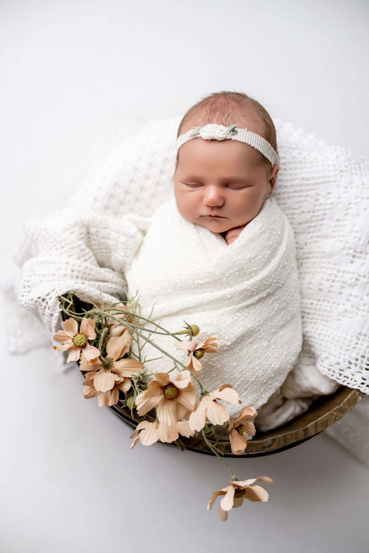 Newborn baby swaddled in white wrap, sleeping in wooden bowl with pink flowers and white headband.