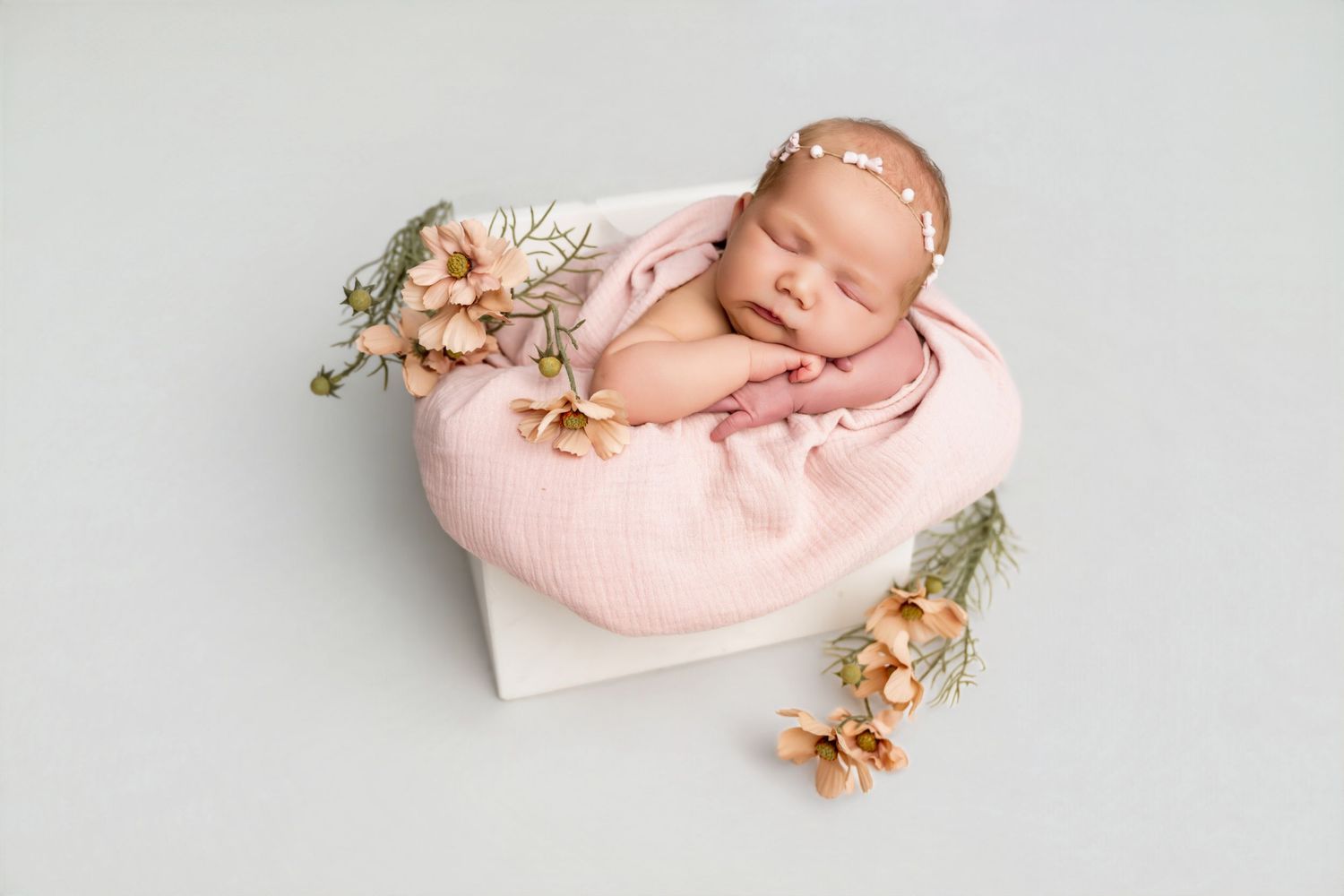 Newborn baby sleeping peacefully in pink bowl with floral accents and delicate headband on gray background.