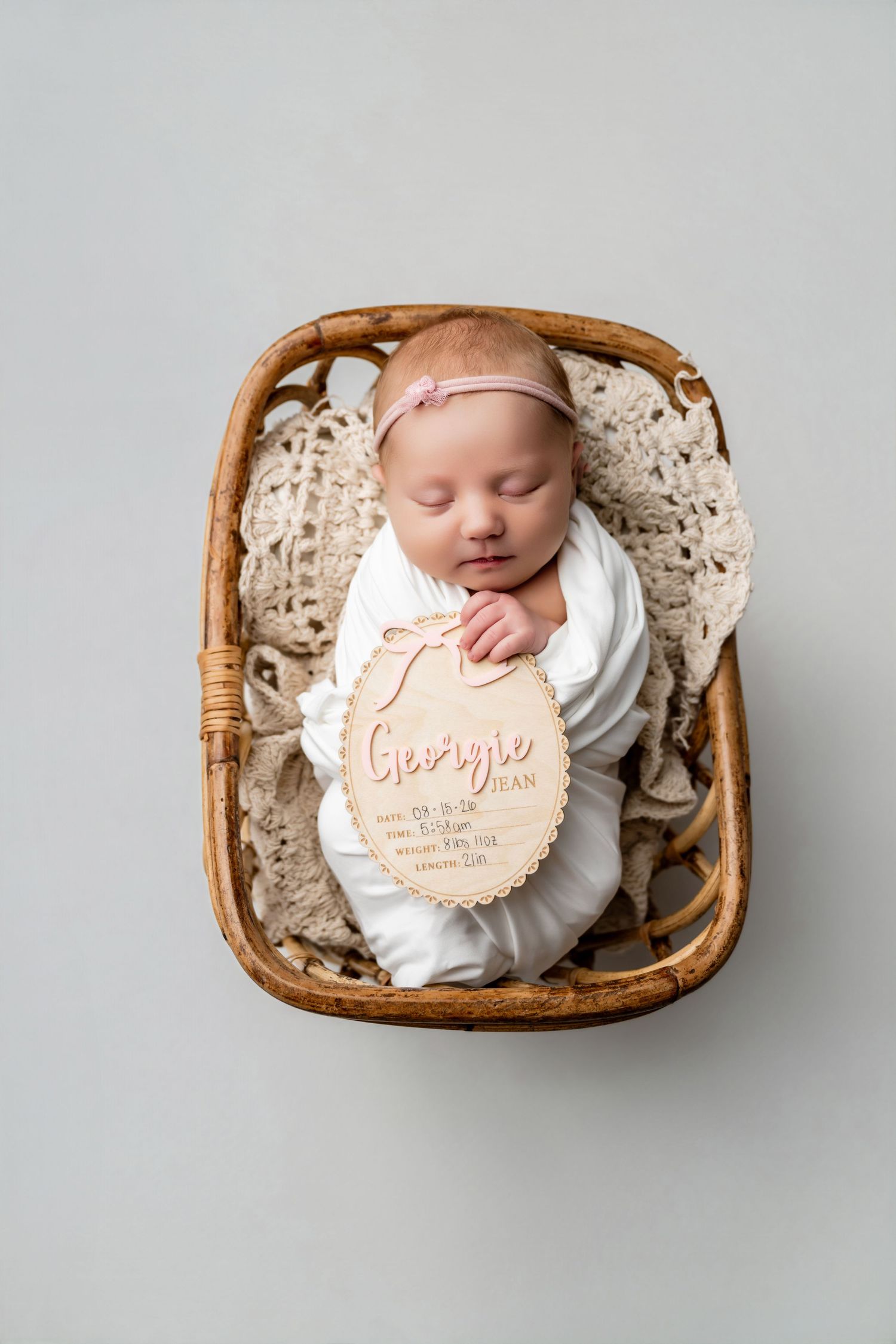 Newborn baby girl named Georgie swaddled in white, sleeping in a wicker basket with a wooden birth announcement sign.
