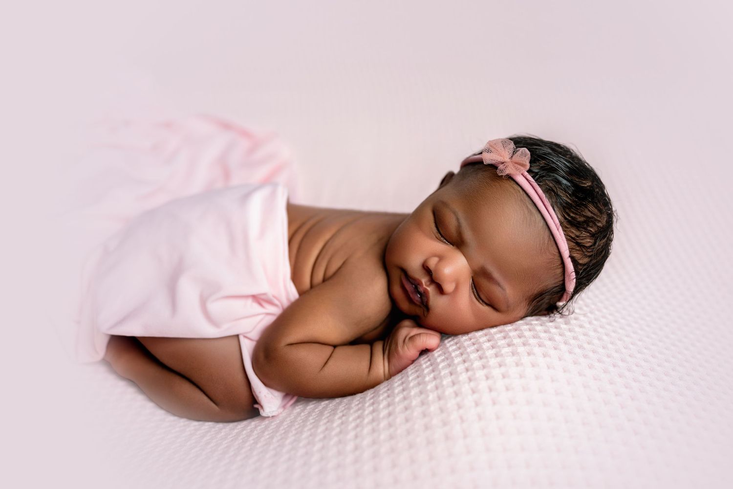 Newborn baby girl sleeping peacefully wrapped in pink blanket with floral headband on soft white background.