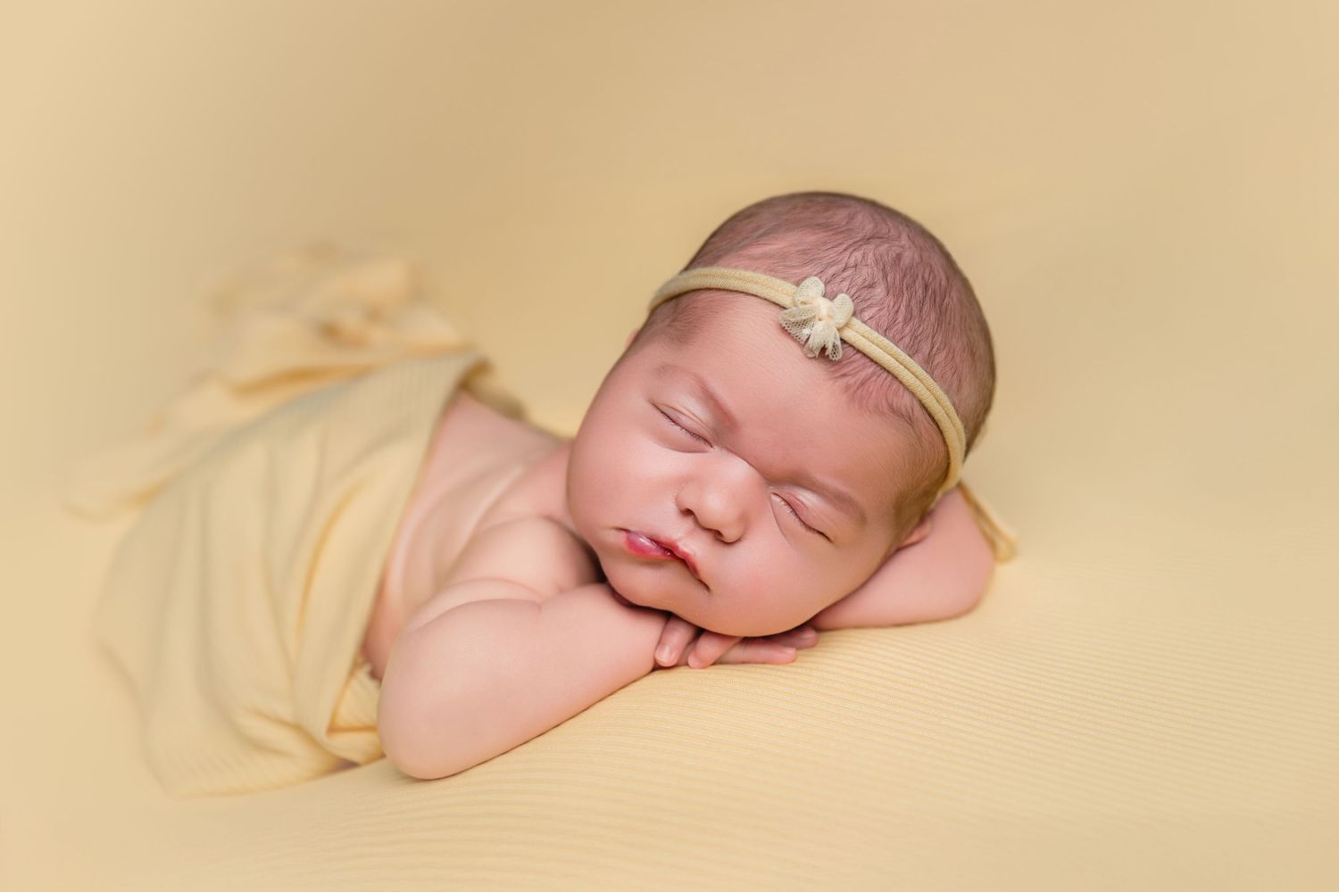 Peaceful newborn baby sleeping on yellow blanket wearing a delicate floral headband in a professional photo.
