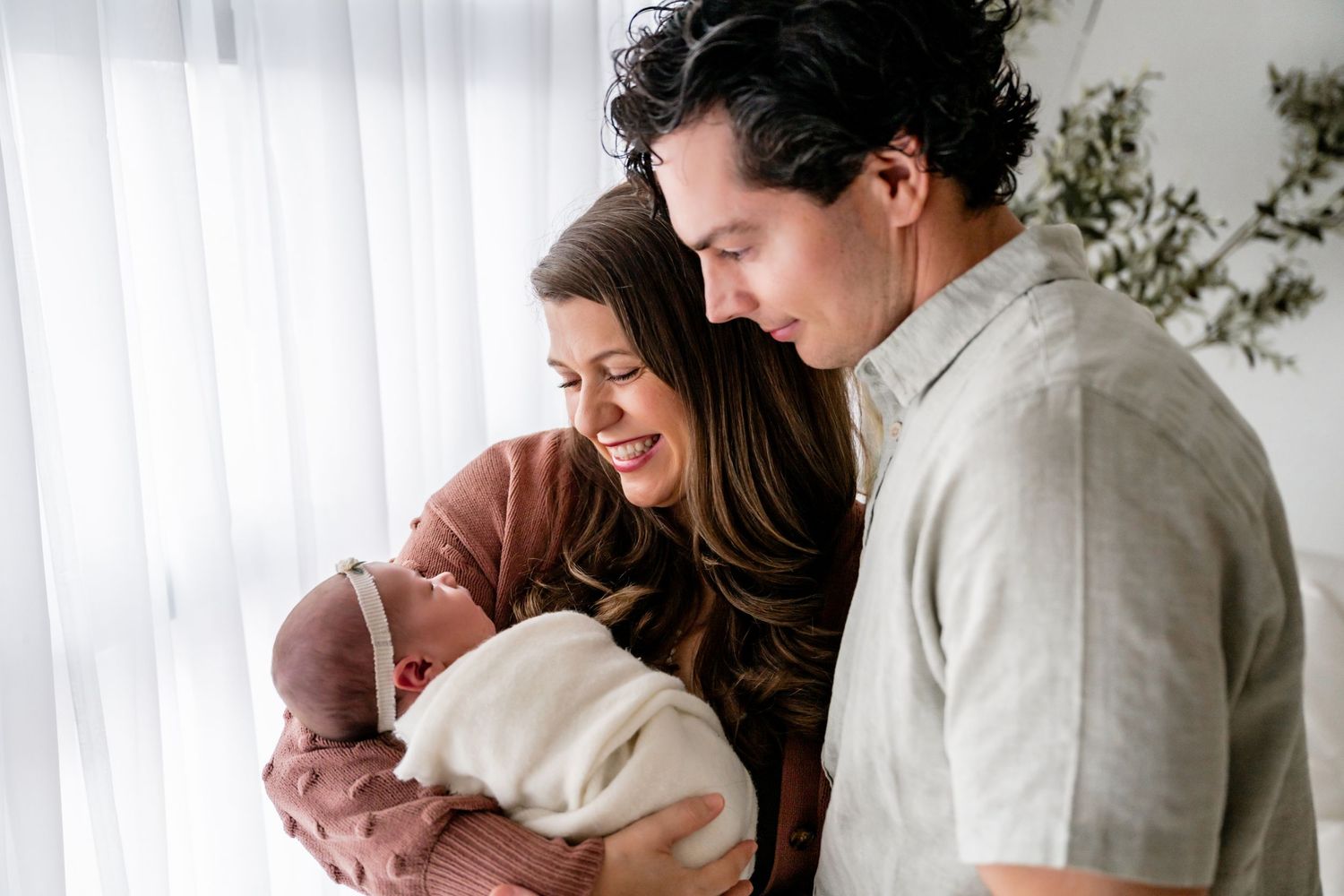 Happy couple smiling and bonding with their newborn baby wrapped in white blanket near bright window.