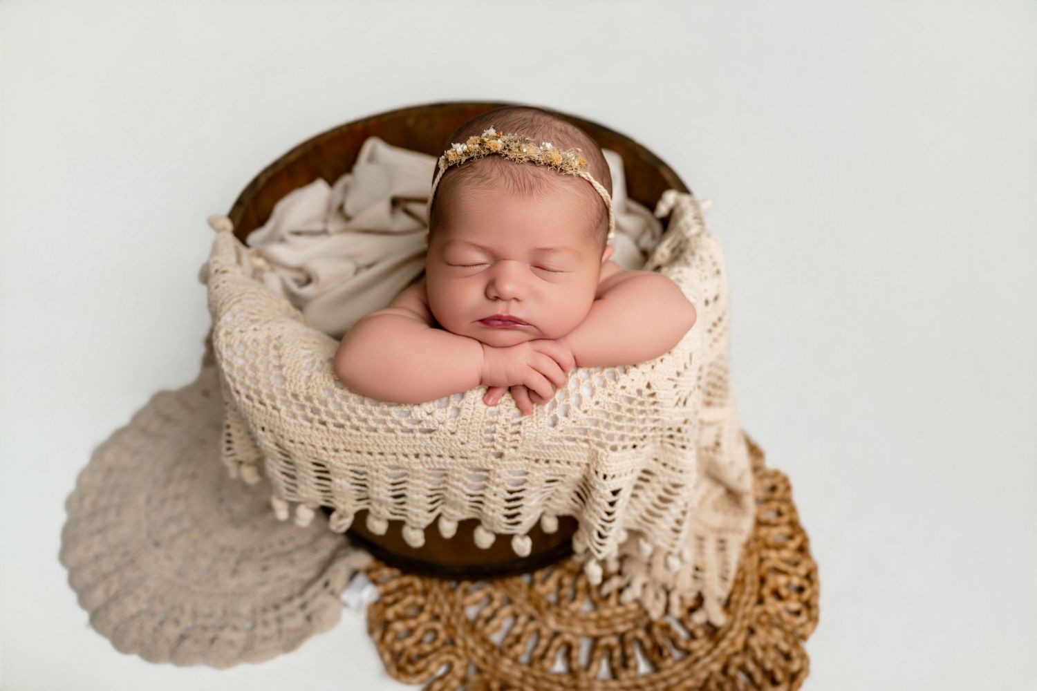 Newborn baby sleeping peacefully in a wooden bowl with cream knit blanket and floral headband.