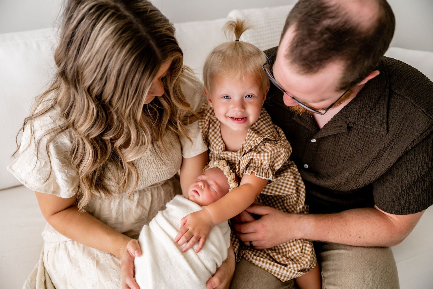 A happy family of four with parents and toddler girl with pigtails smiling at their newborn baby wrapped in white.