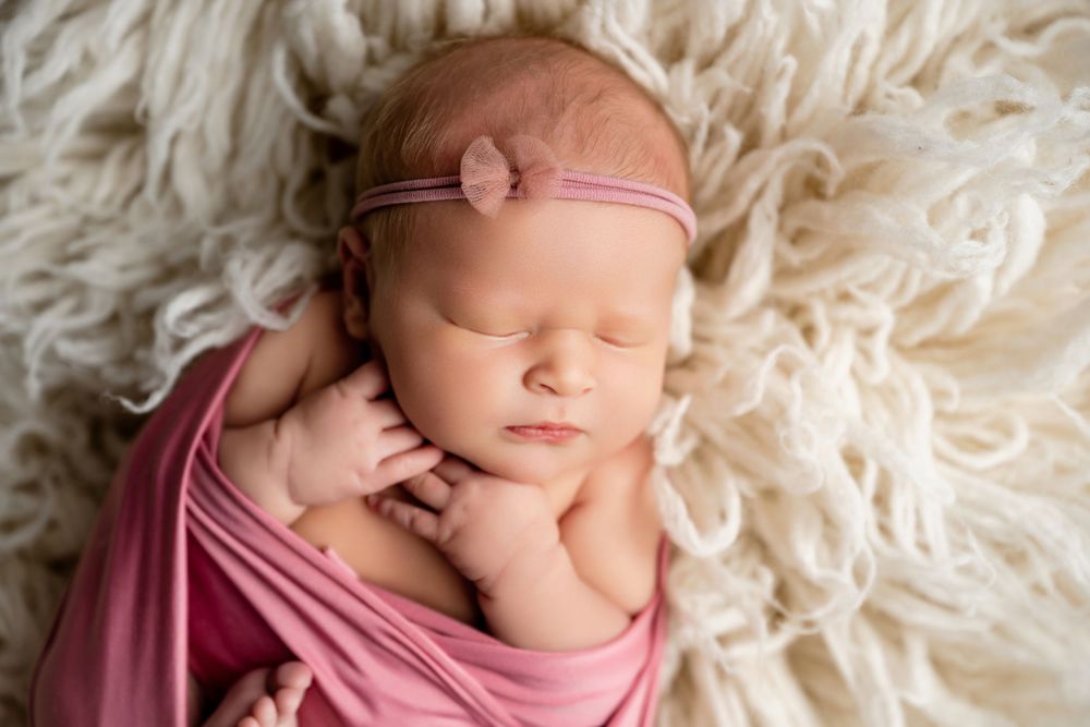 Sleeping newborn baby girl wrapped in pink swaddle with floral headband on fluffy white fur backdrop.