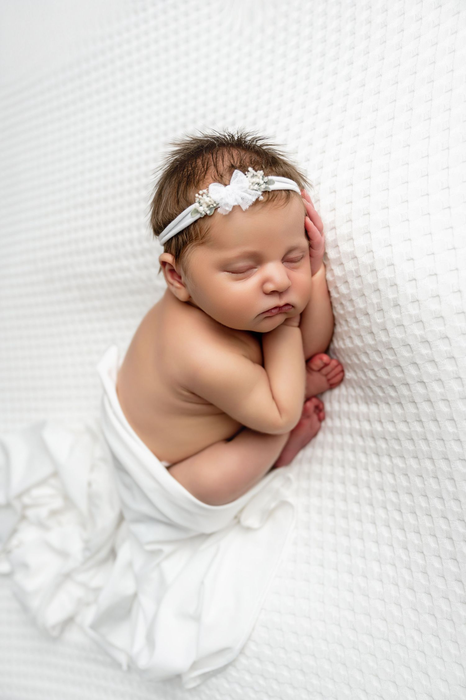 Sleeping newborn baby girl wearing a white floral headband, wrapped in a white blanket on a white textured background.