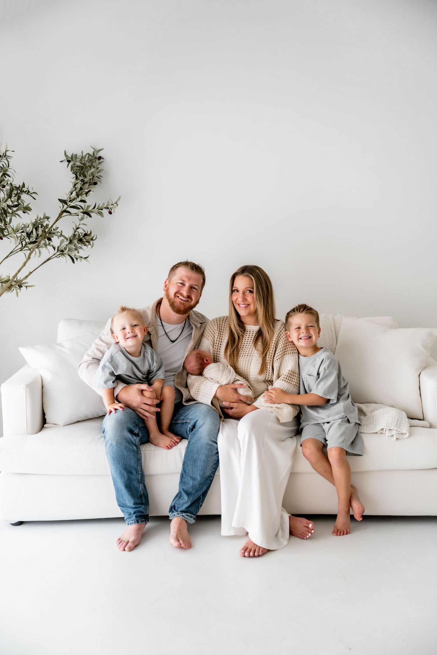Happy family of five with newborn baby posing on white couch in bright minimalist studio portrait.