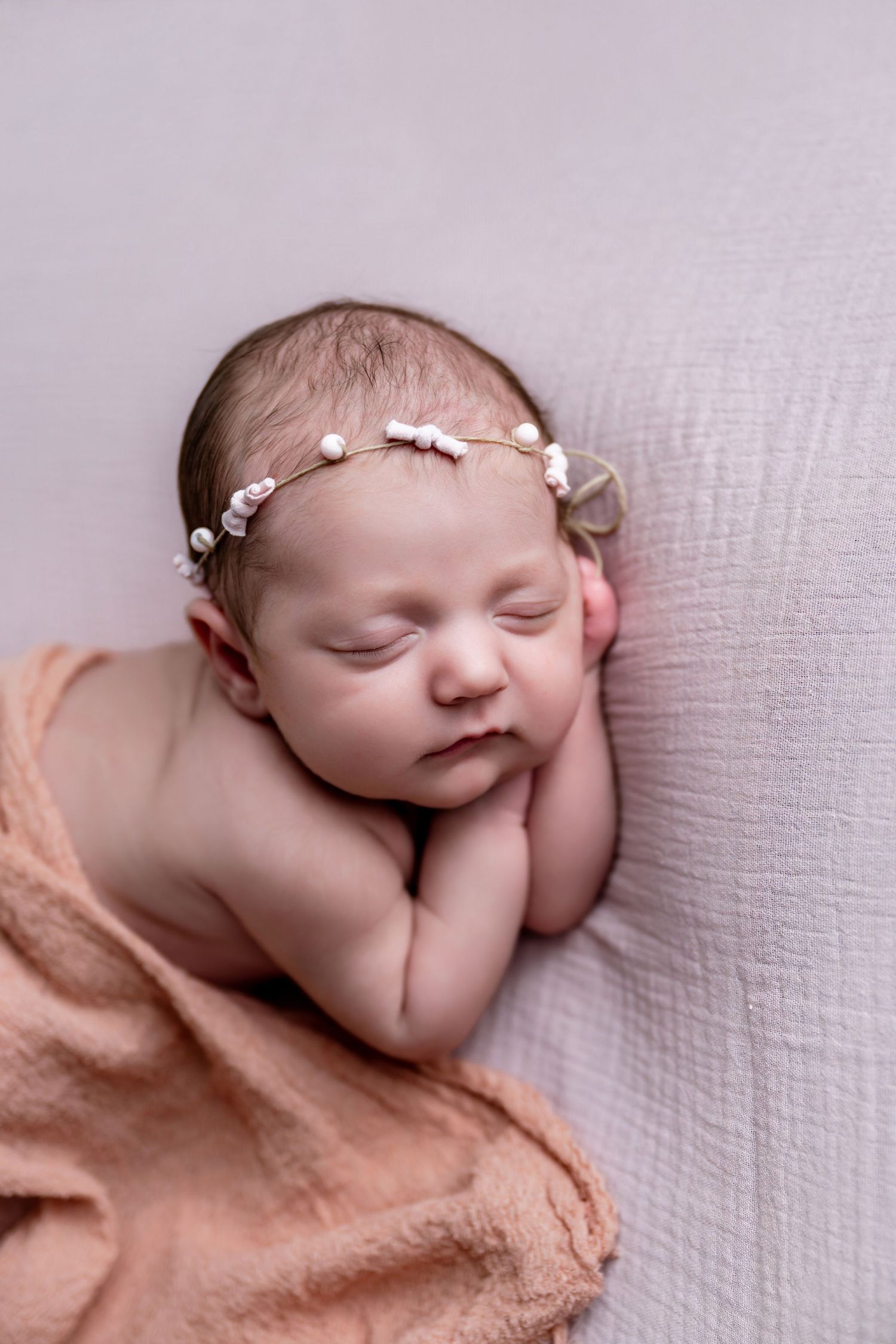 Peaceful newborn baby sleeping with hands under chin, wearing a delicate floral headband, wrapped in a soft peach blanket.