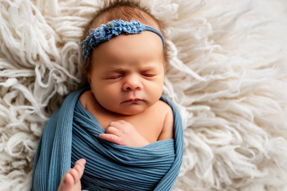 Newborn baby wrapped in soft blue swaddle cloth sleeping peacefully on white fluffy rug.