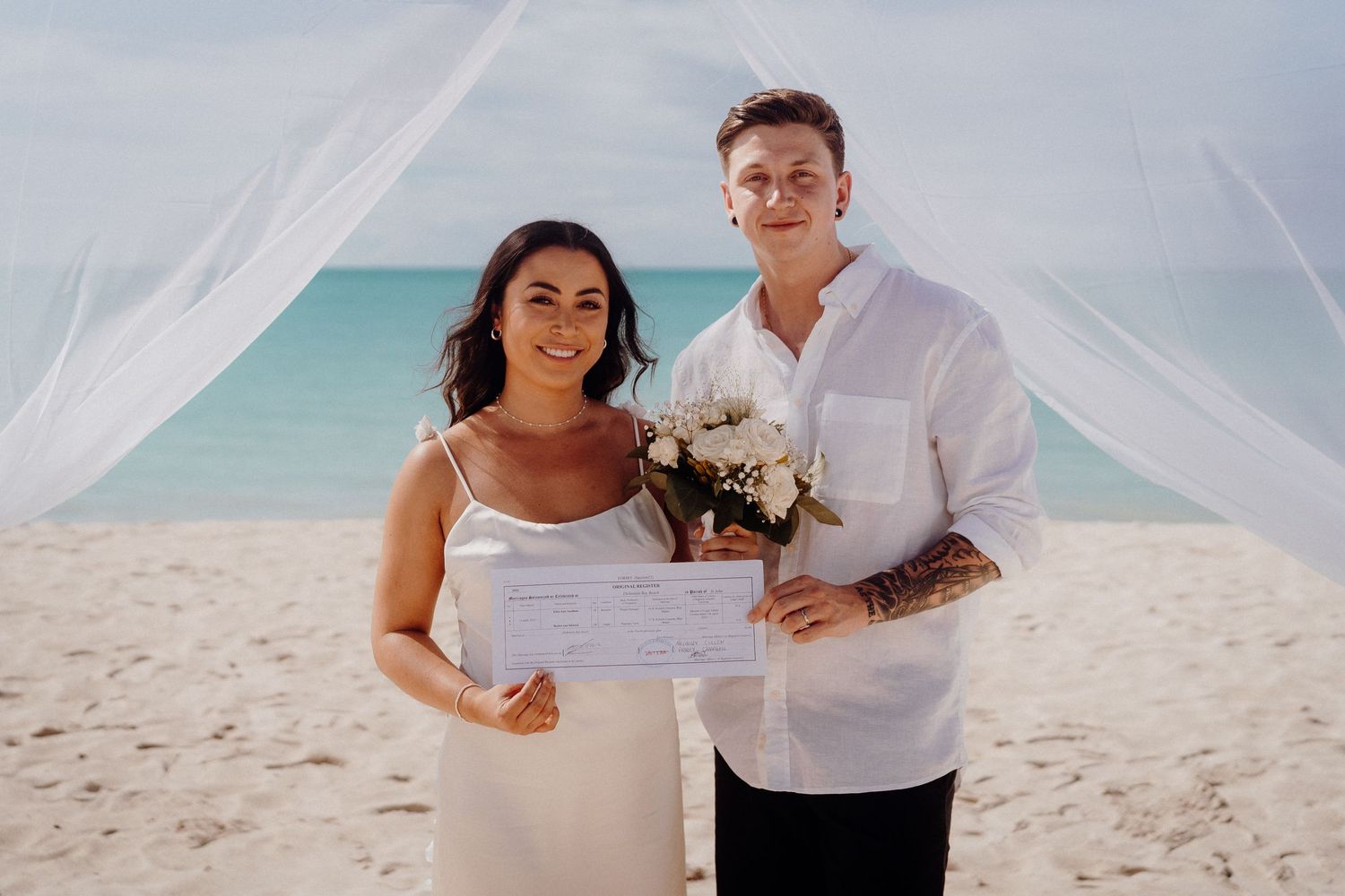Beach wedding ceremony with couple under white fabric canopy against turquoise waters.