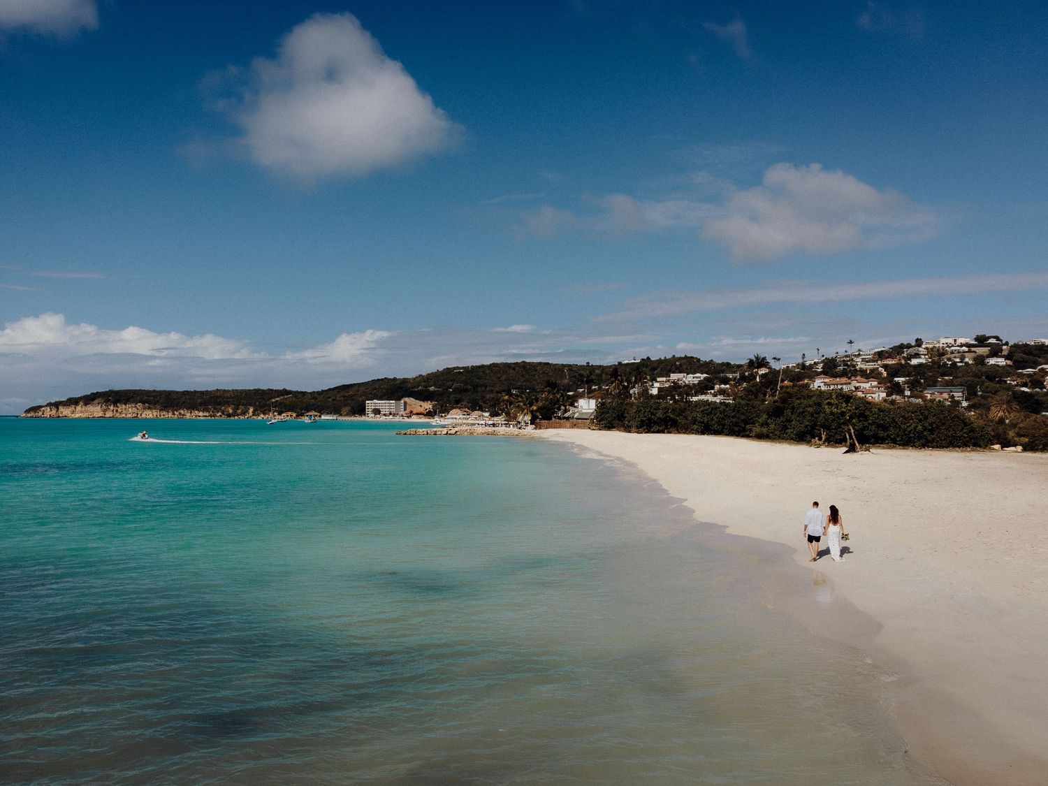 Beautiful turquoise beach with white sand and houses on hillside under blue sky with puffy clouds.