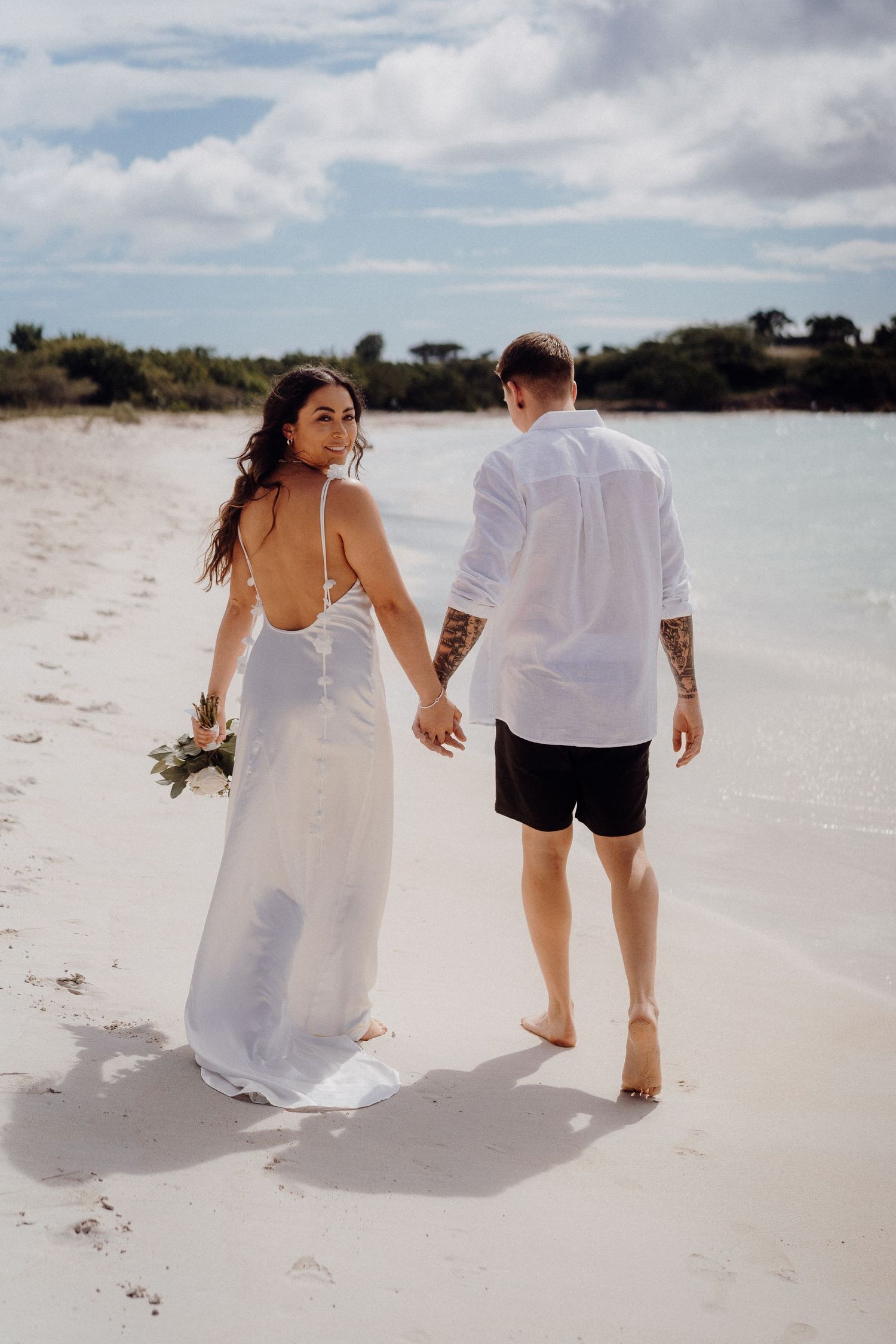 Newlyweds walking hand in hand on pristine white sand beach.