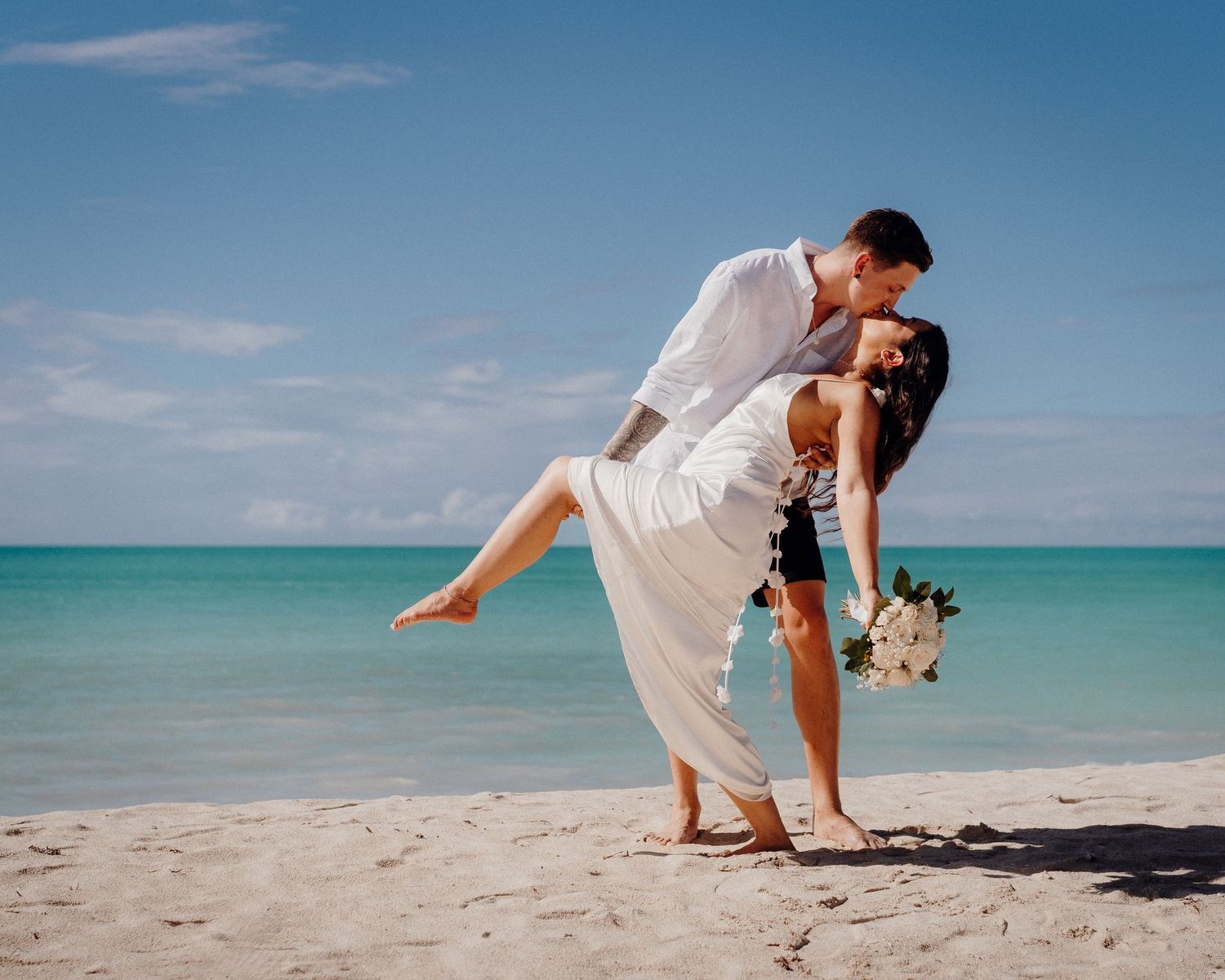 Romantic beach photoshoot with dramatic dip pose against turquoise ocean backdrop.