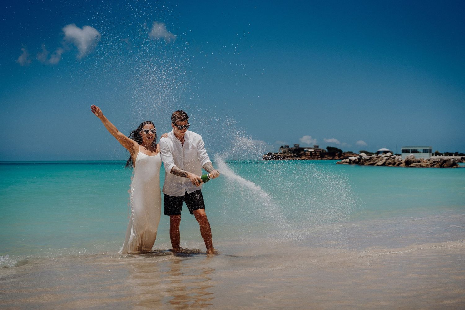 Playful couple celebrating on beach with water splashes and vibrant turquoise ocean view.