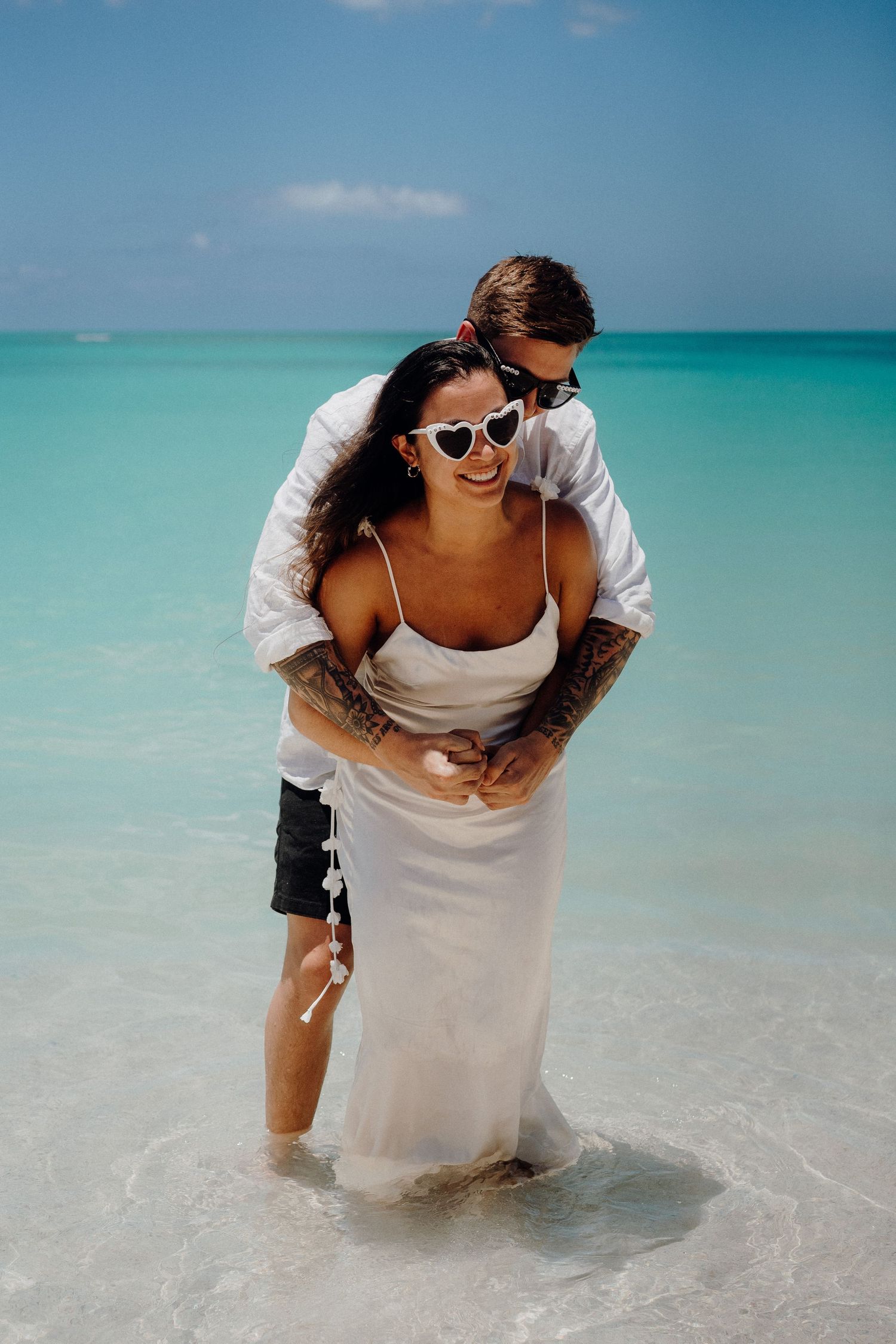 Romantic beachside moment with couple embracing in white attire against turquoise ocean backdrop.