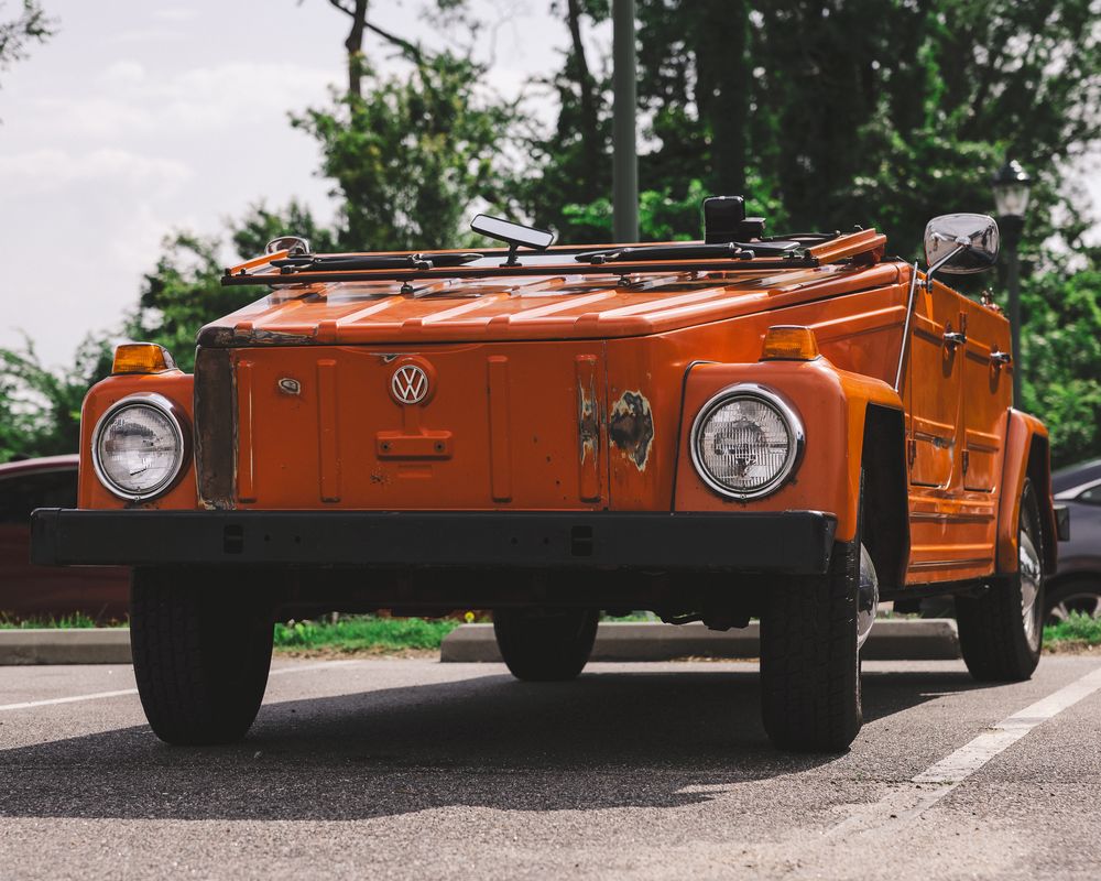 Vintage orange Volkswagen Thing 181 off-road vehicle driving on tree-lined road with roof rack.