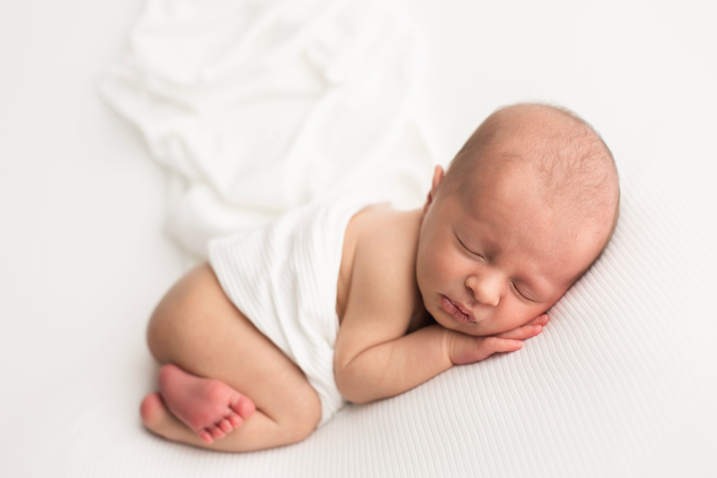Sleeping newborn baby in white wrap against light background during professional portrait session.