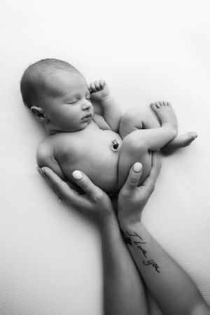 Artistic black and white portrait of newborn baby supported by tattooed arms on white background.