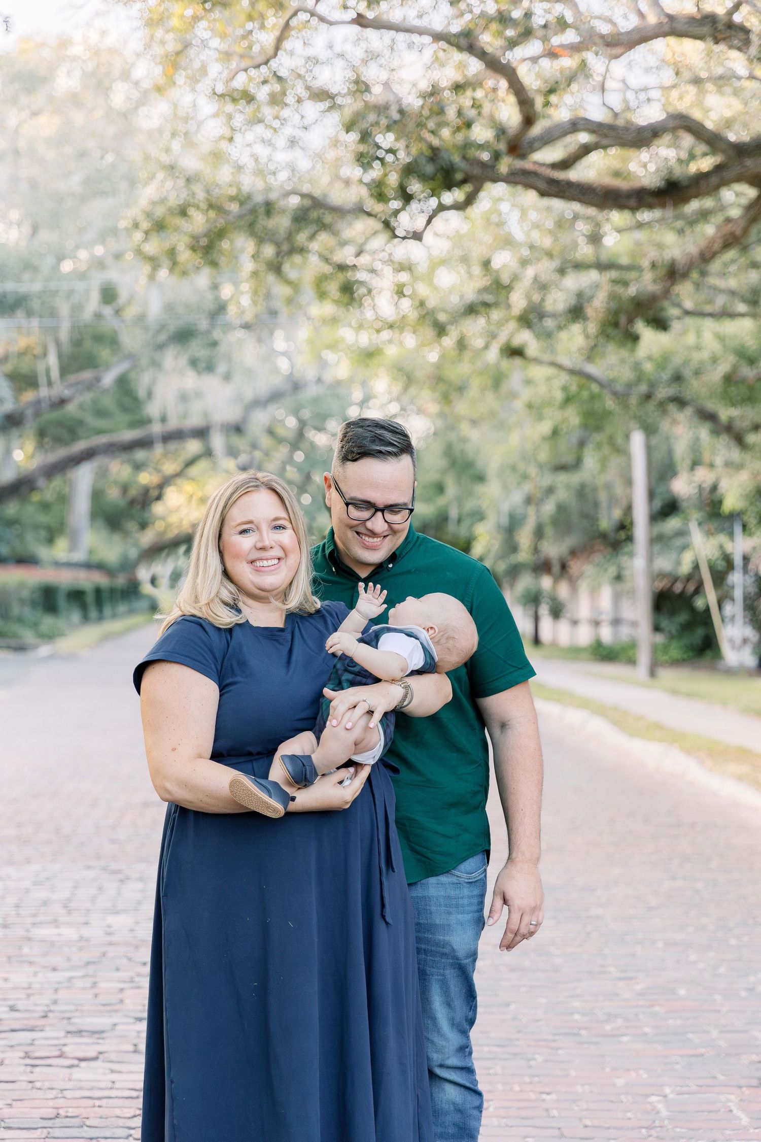 A couple poses outdoors with their baby under a canopy of trees.