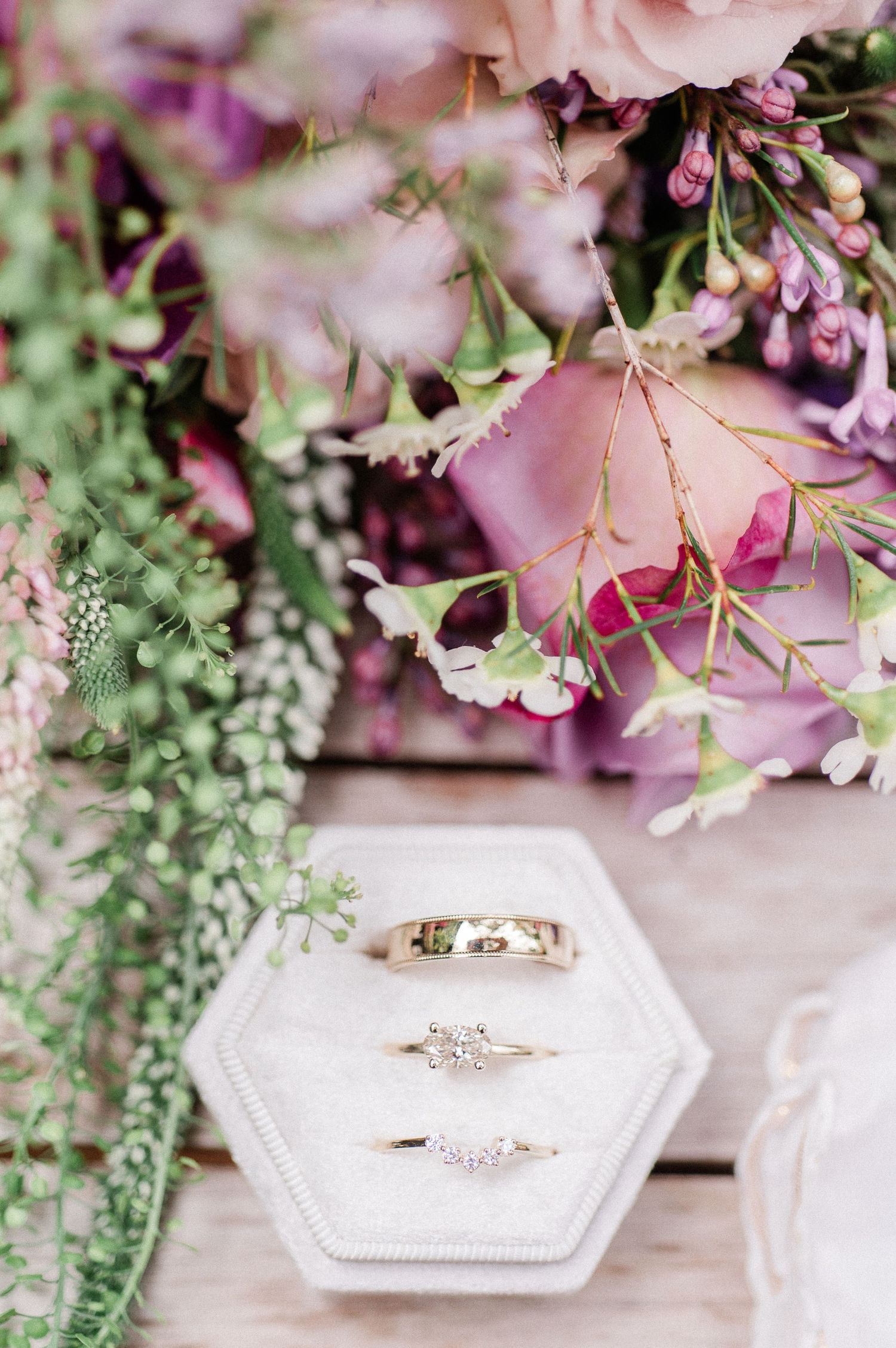Wedding rings displayed on white hexagonal box surrounded by delicate pink and purple floral arrangements.