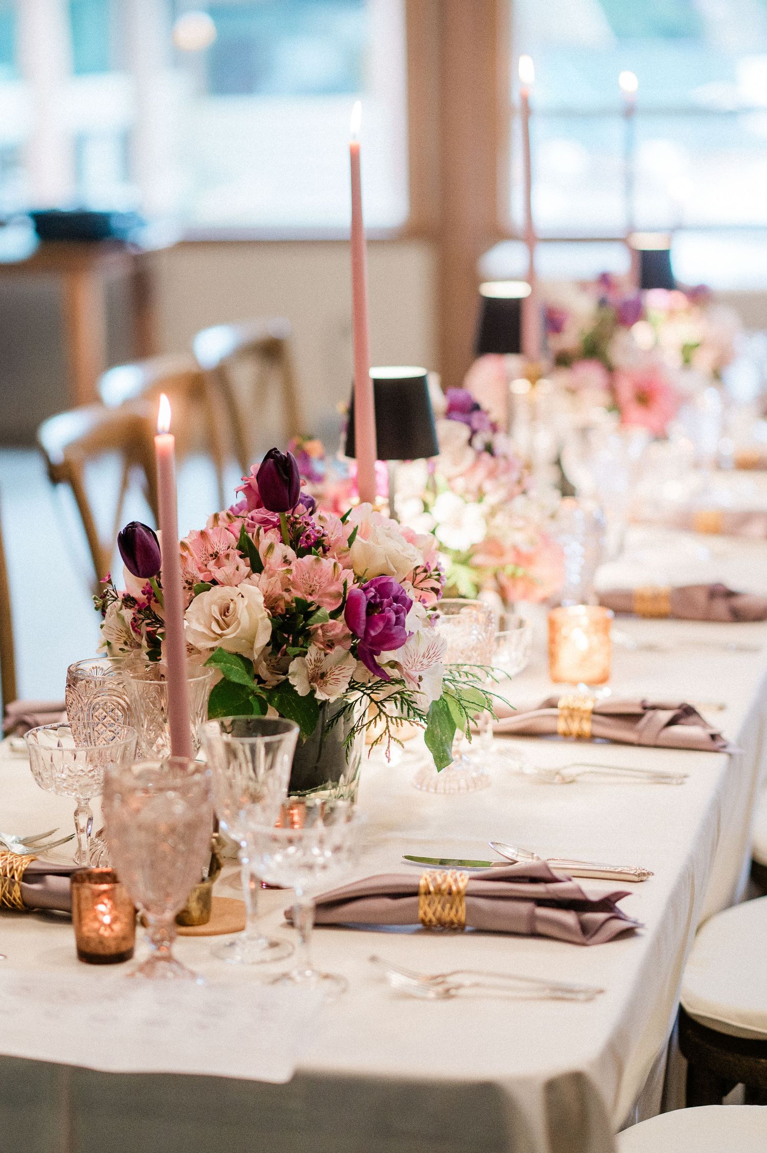 Elegant wedding table setting with pink and purple floral centerpieces, candles, and crystal glassware on white linens.