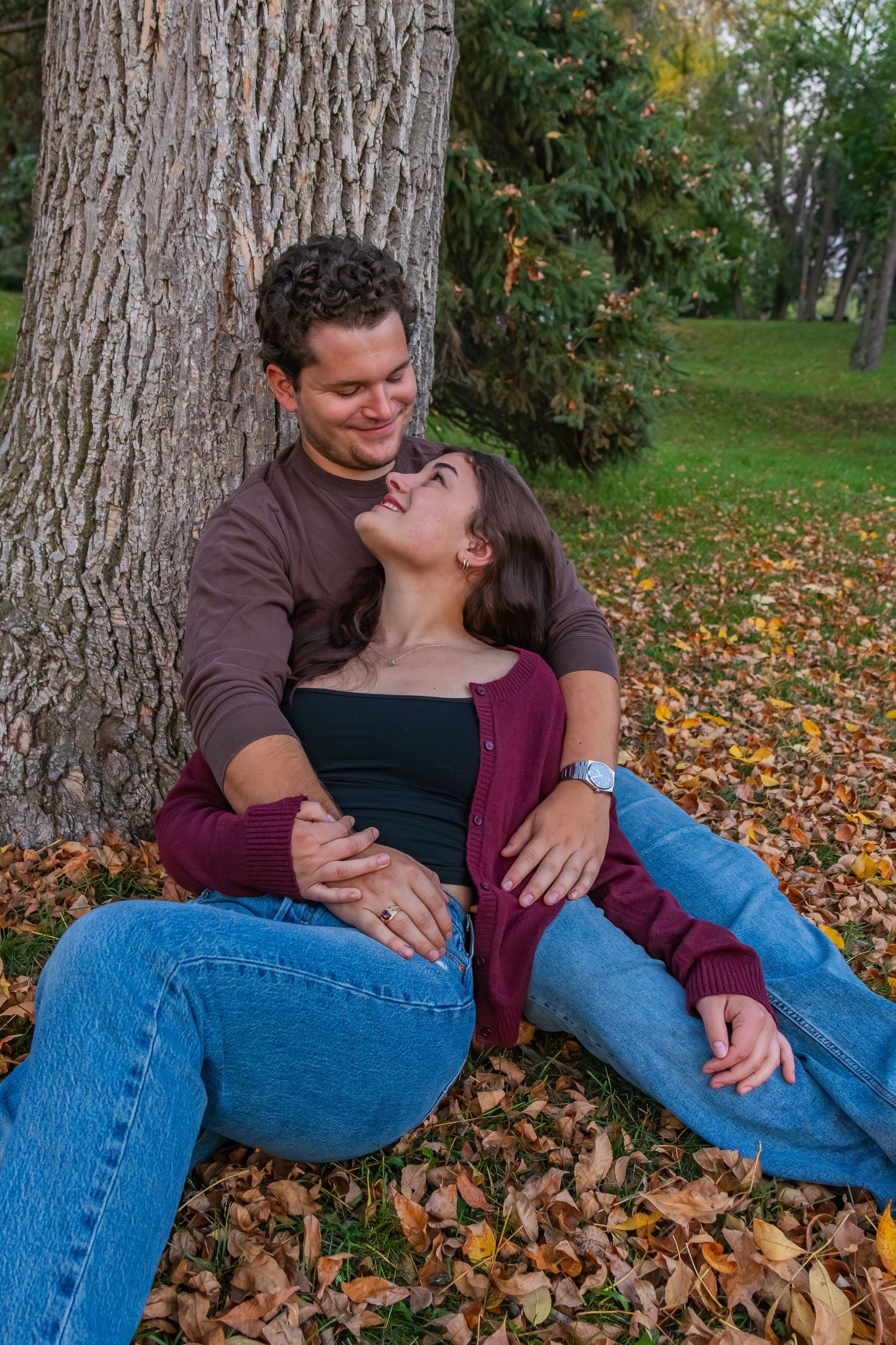 Couple sitting together against a tree surrounded by colourful autumn leaves in a park setting. Blue jeans