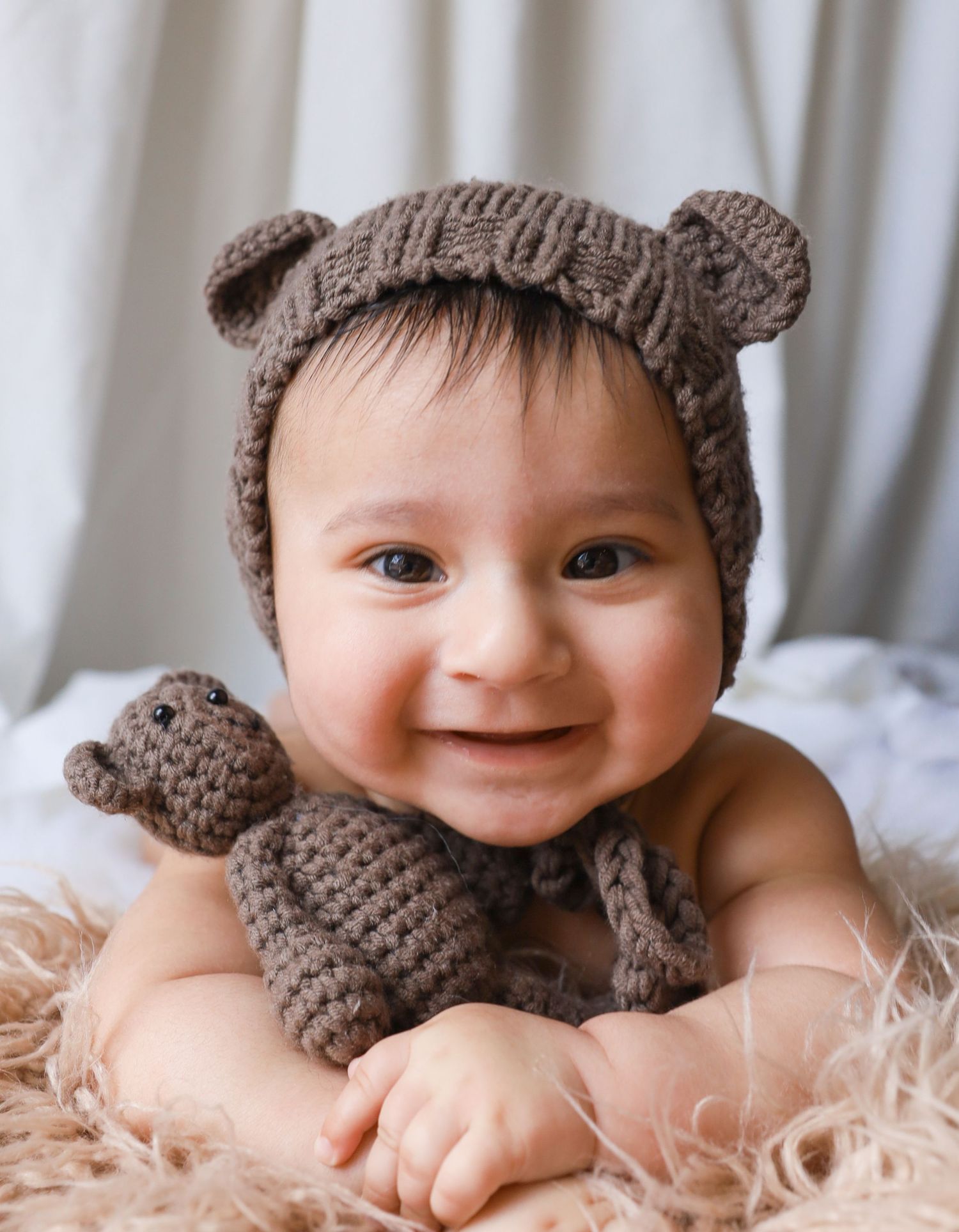 Baby wearing knitted brown bear hat and matching toy on furry blanket.