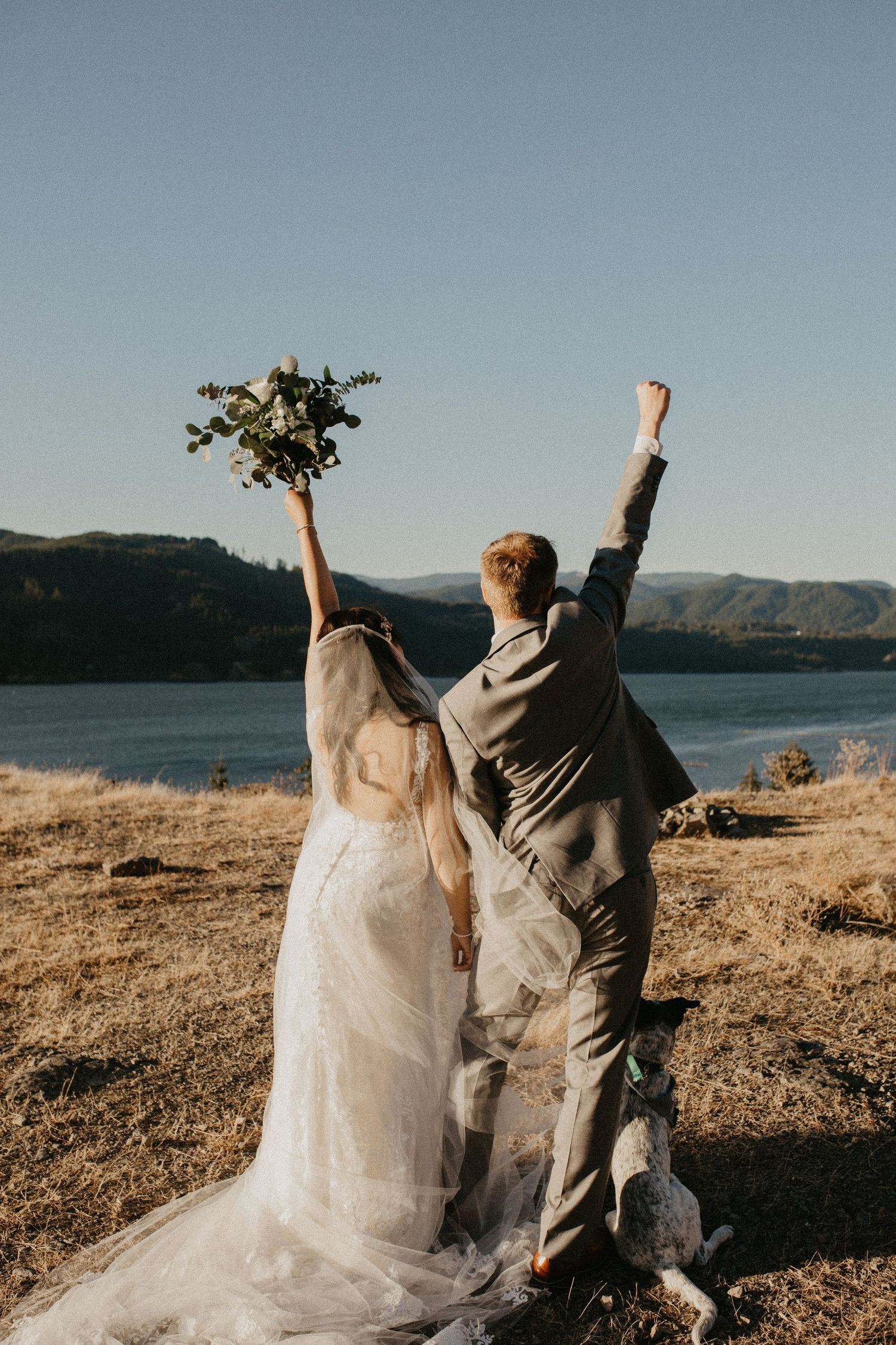 Newlywed couple celebrating with raised arms against scenic mountain lake landscape at sunset.