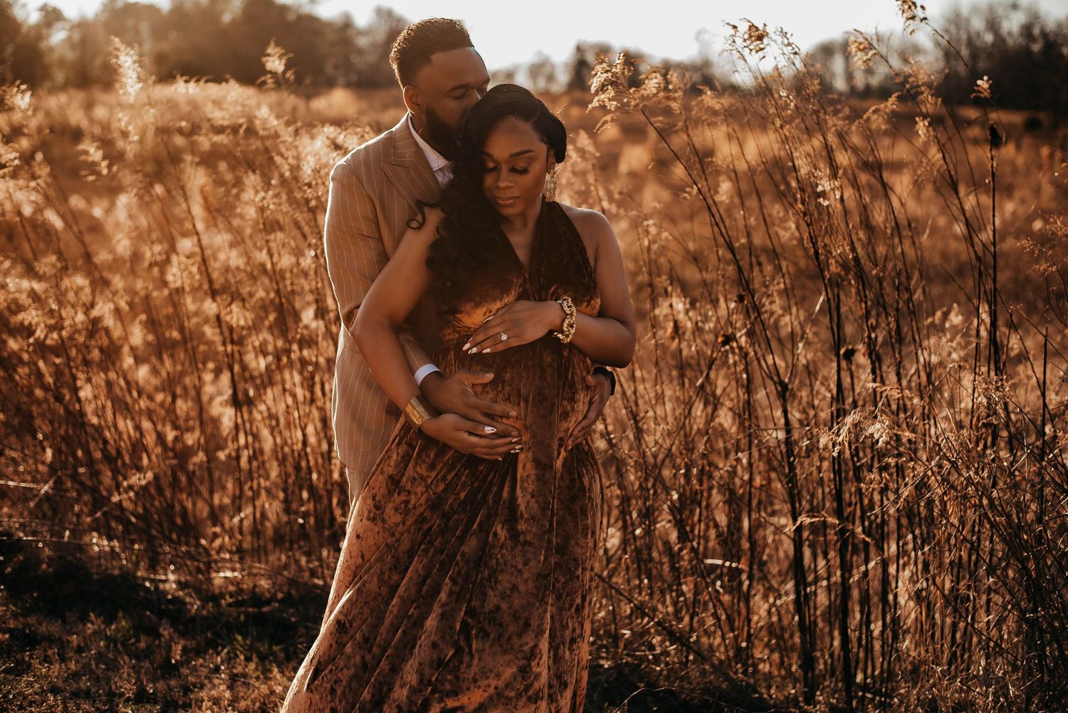 Couple embracing in golden field at sunset, woman in floral maternity dress holding baby bump.