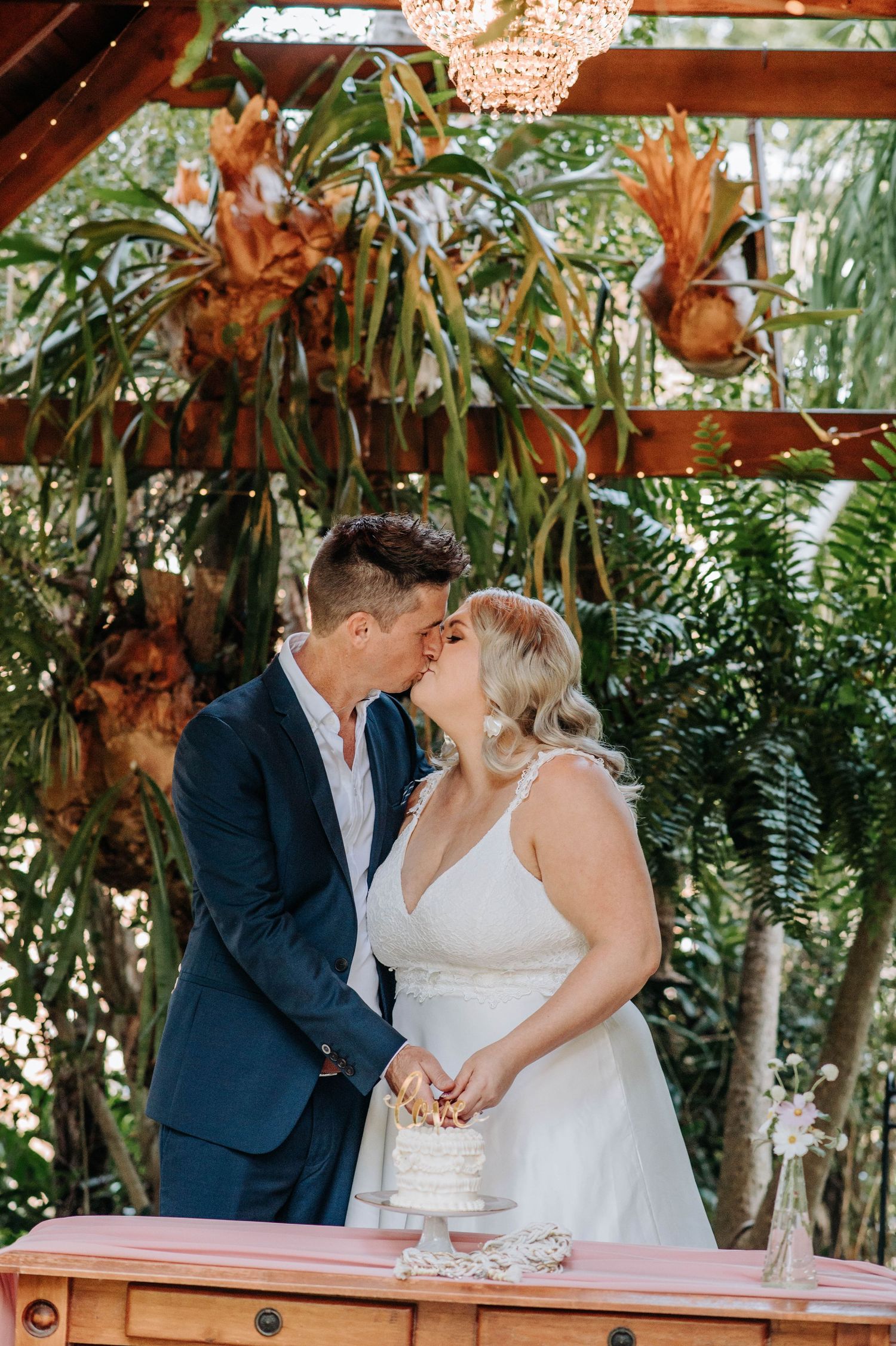 A couple shares an intimate kiss during their wedding ceremony under a wooden pergola decorated with greenery.