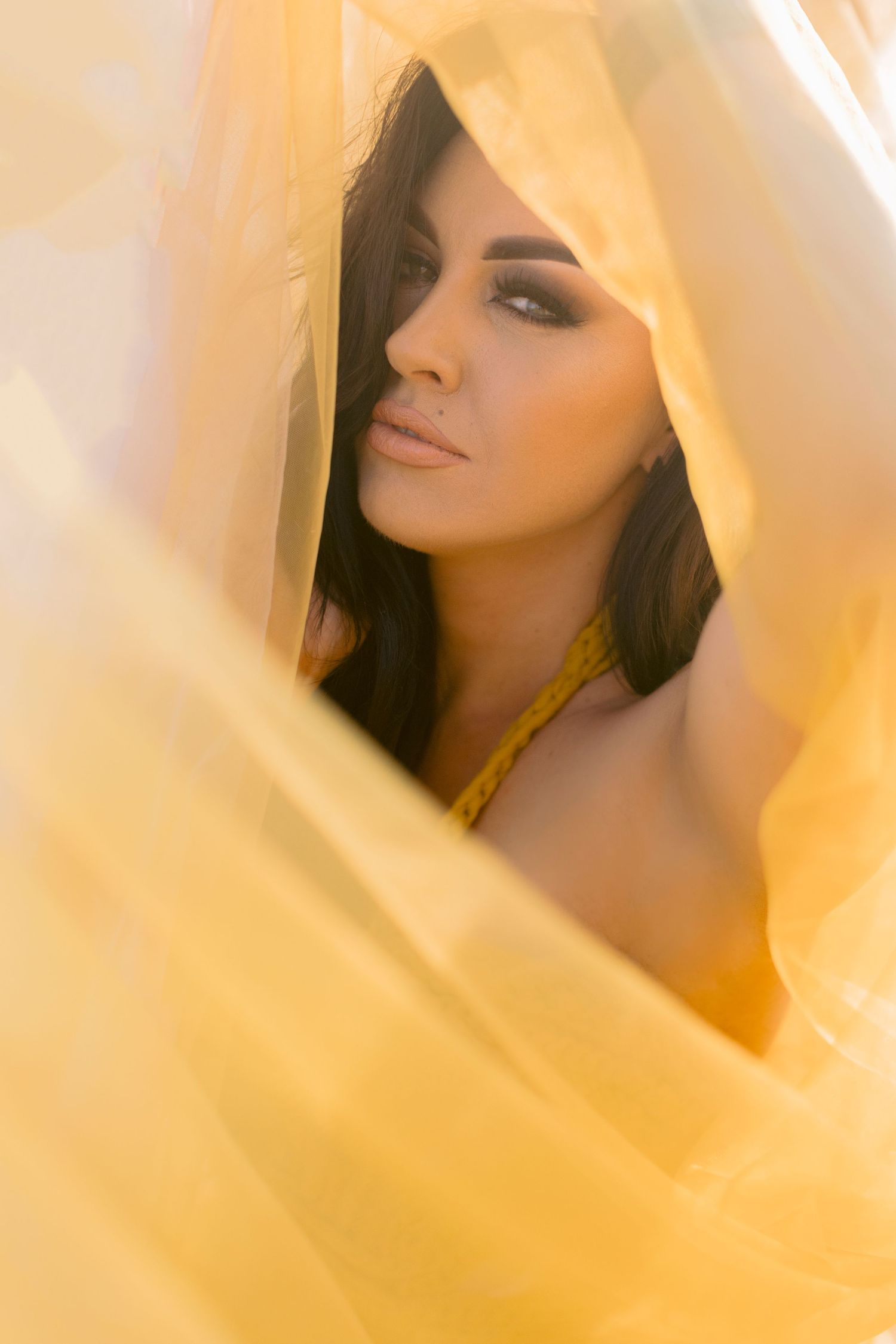 Mysterious brunette woman with dramatic makeup peering through golden yellow fabric.
