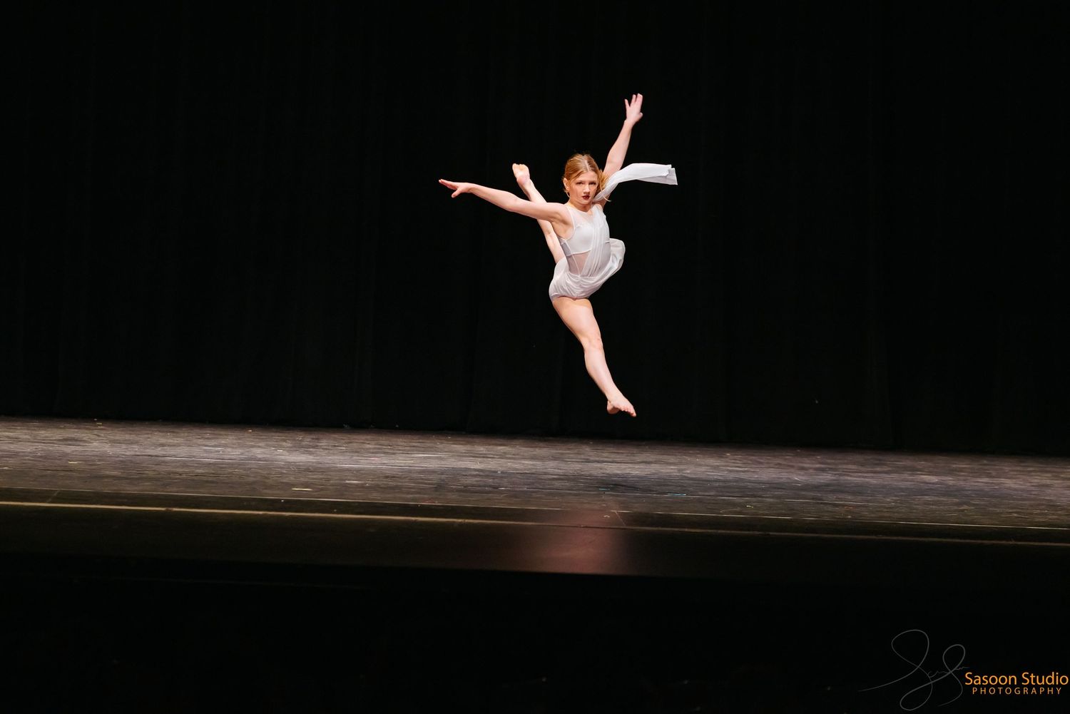 Graceful dancer in white costume leaps across stage during performance.