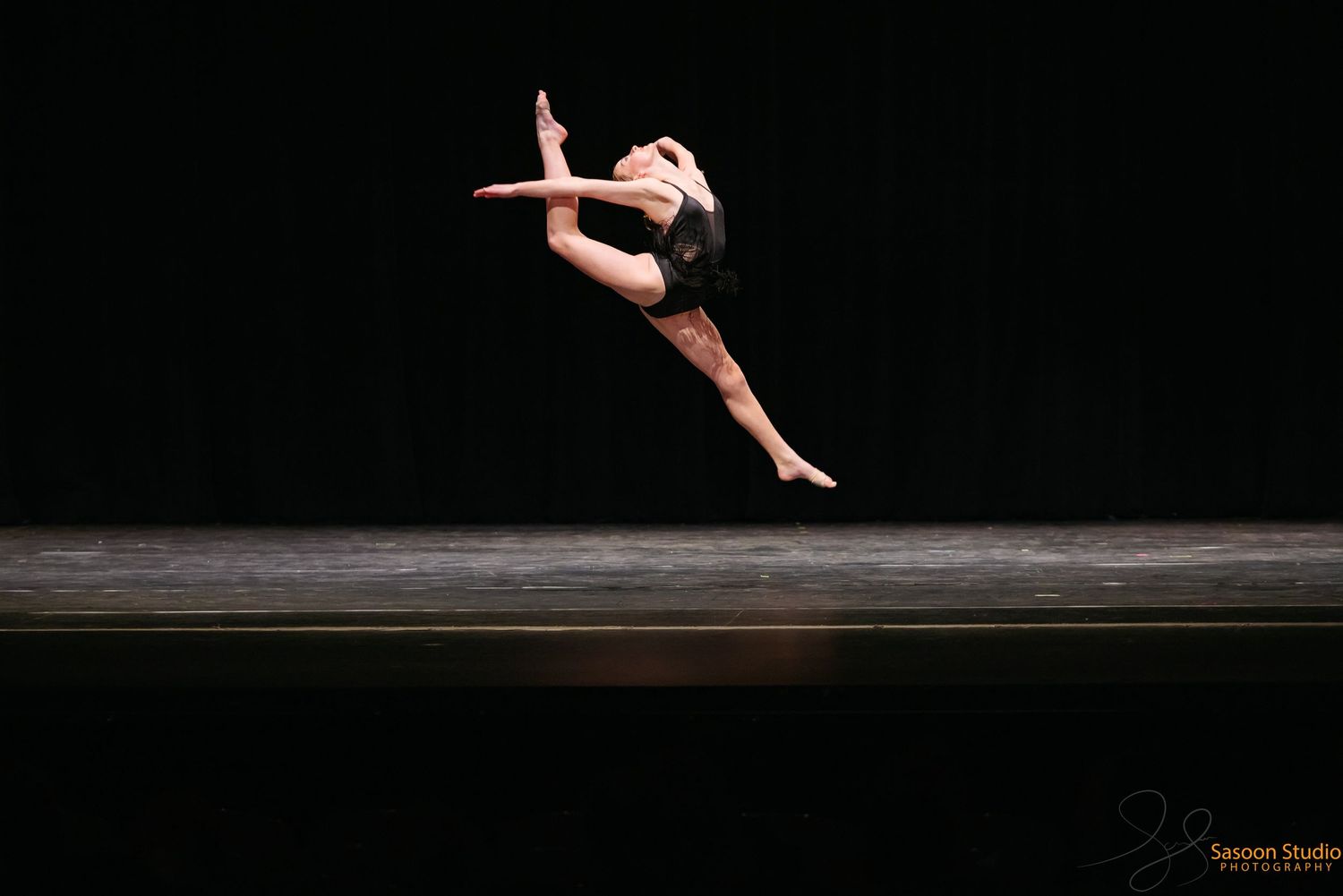 Dancer in black leaps gracefully through the air during a stage performance against dark backdrop.