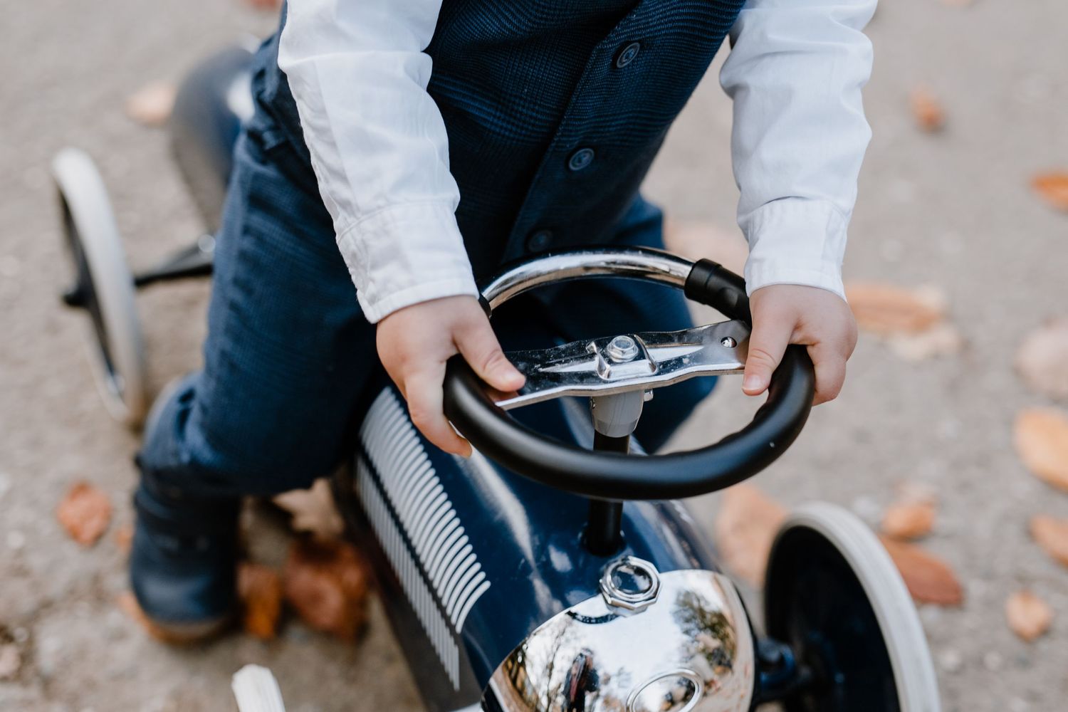 Child riding vintage pedal car with chrome details among fallen autumn leaves.
