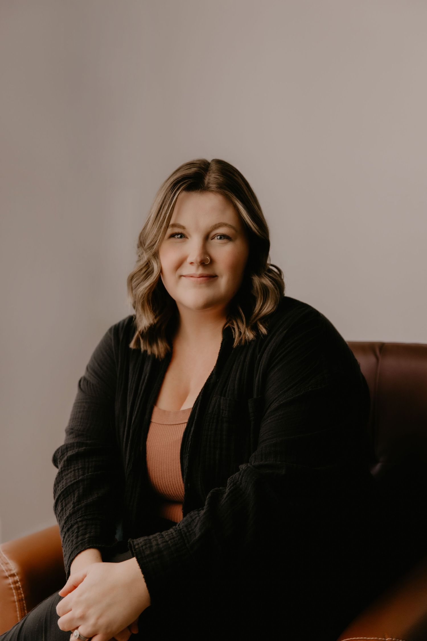 Woman in black cardigan sitting in a brown leather chair, smiling warmly against a neutral gray background.