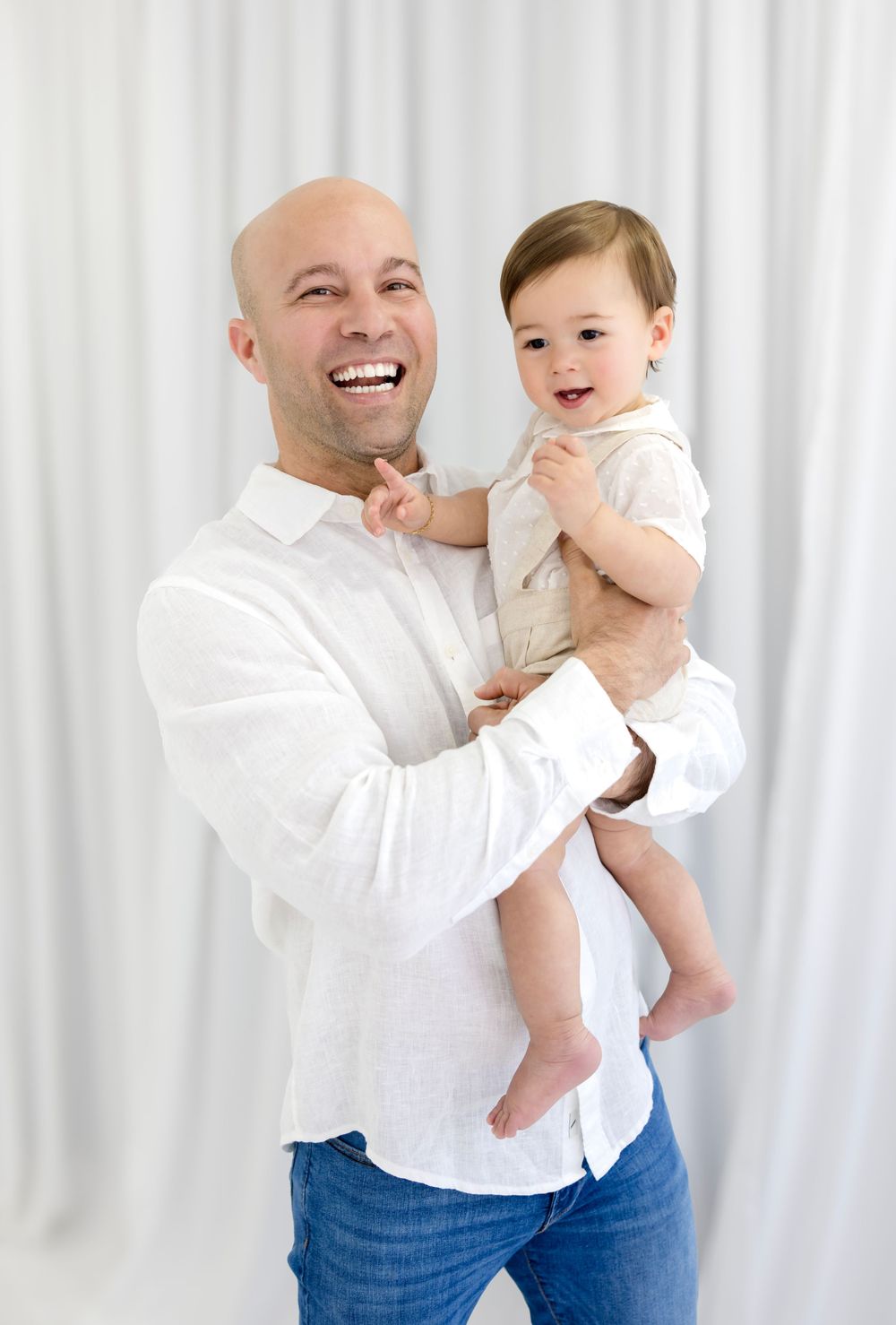 Smiling bald man in white shirt holding a happy baby in a bright white studio setting.