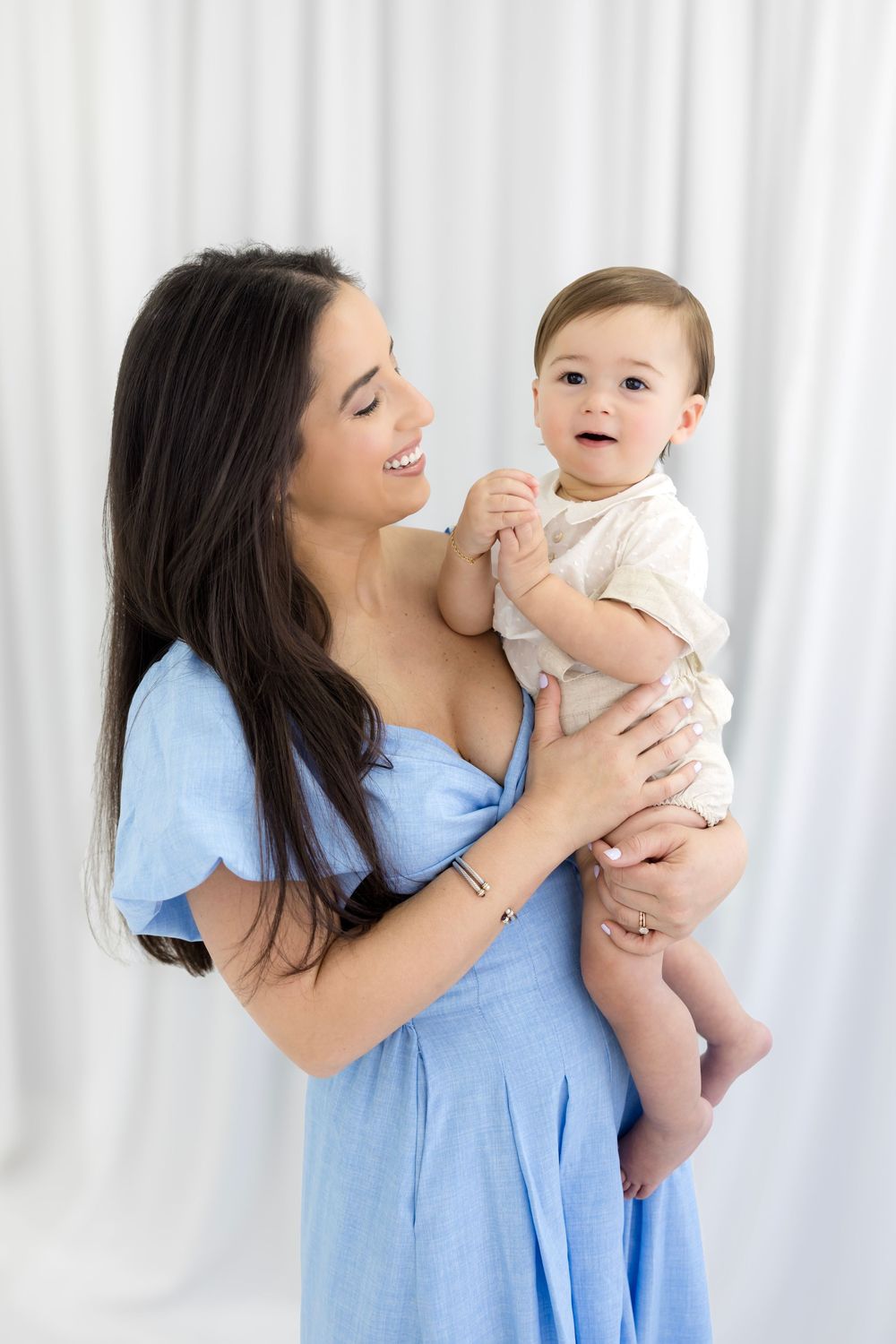 Smiling brunette woman in blue dress lovingly holds a happy baby in a white romper against a white backdrop.
