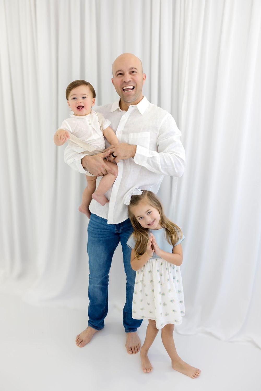 Bald father laughing while holding baby son and standing with smiling daughter in white studio photo.