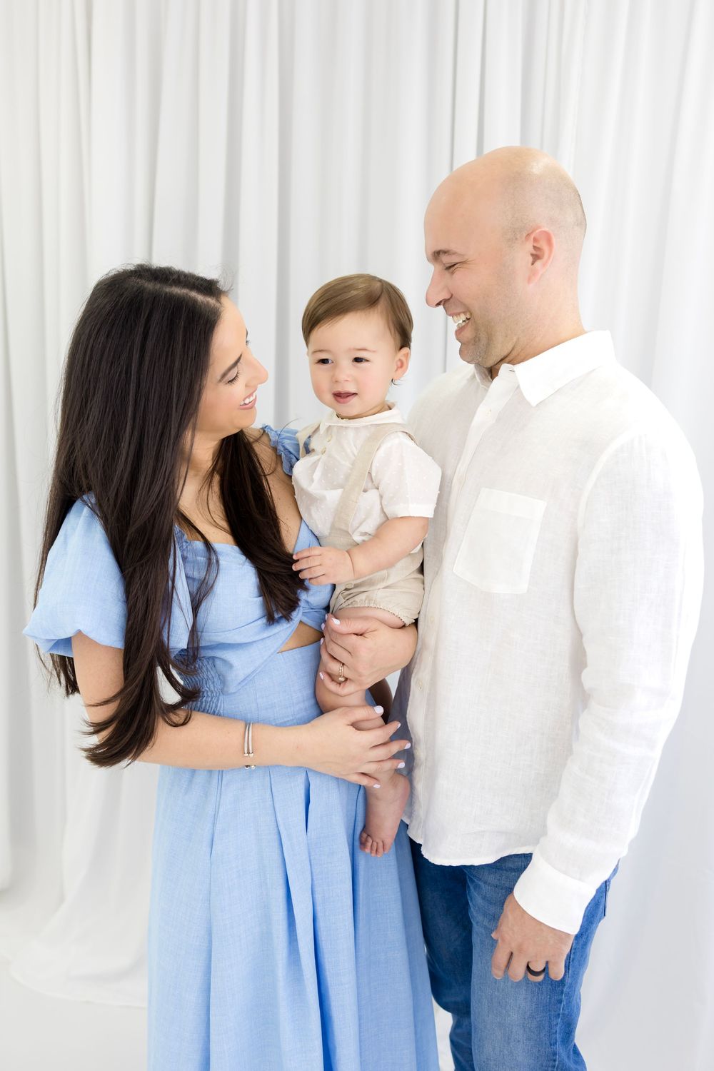 A happy family with a bald father, dark-haired mother in blue dress, and smiling baby boy in a white studio setting.