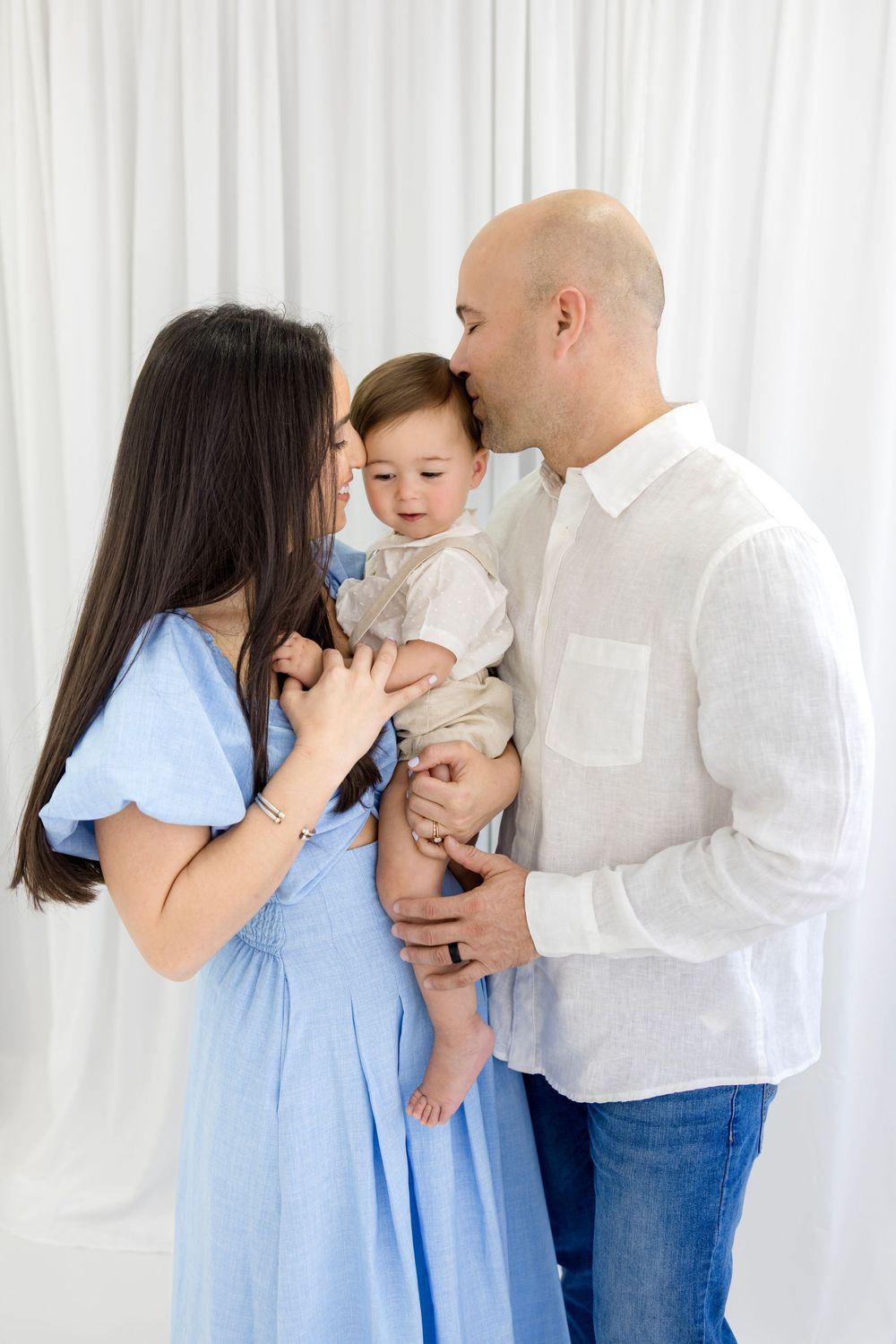 A loving family portrait with parents holding their baby boy against a white backdrop.