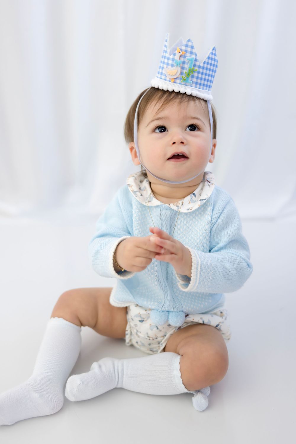 Cute baby wearing a blue gingham crown, light blue cardigan, and white knee socks, sitting on a white background.