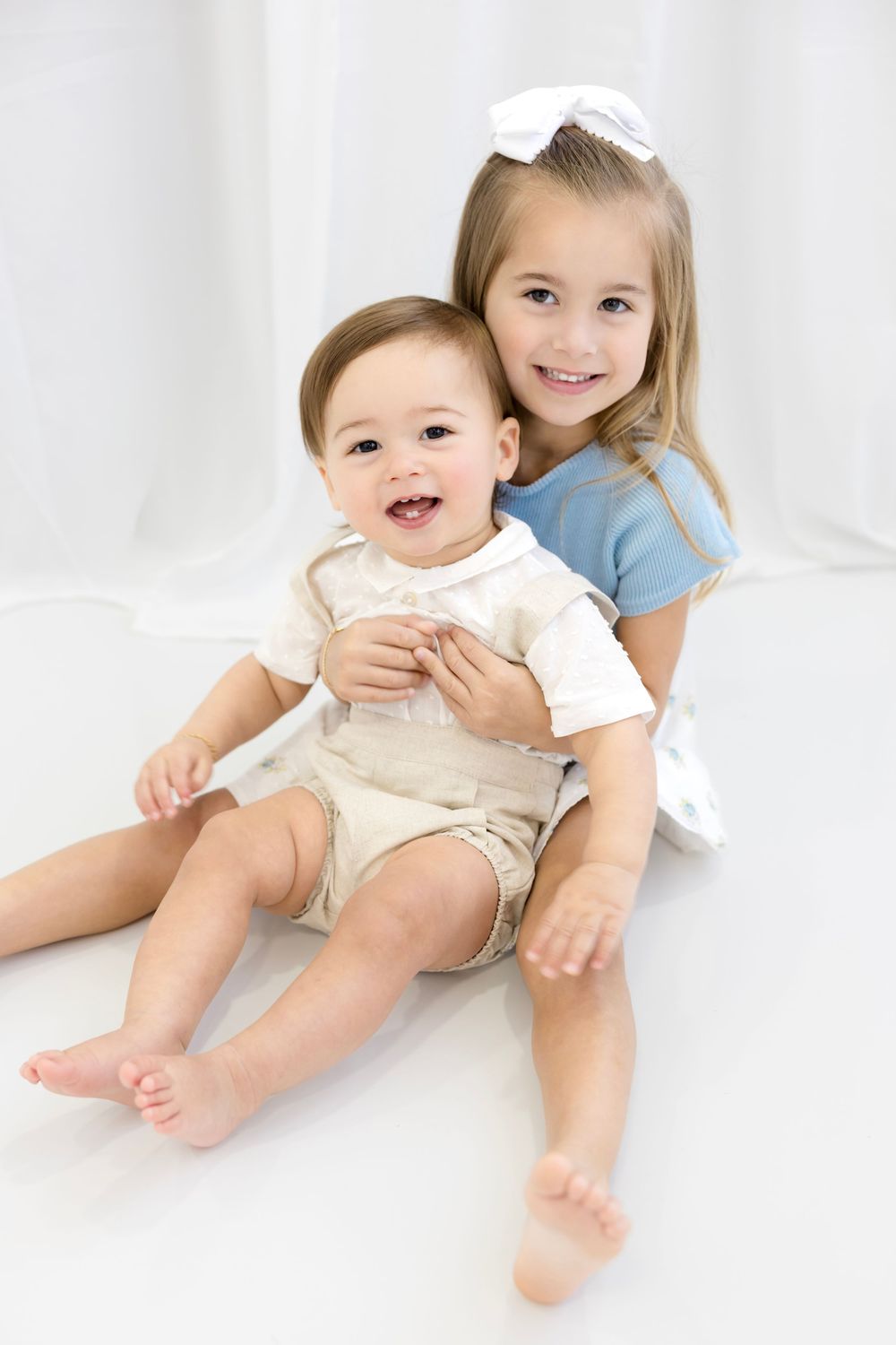 A smiling baby boy and older sister with a white bow pose together on a white background.