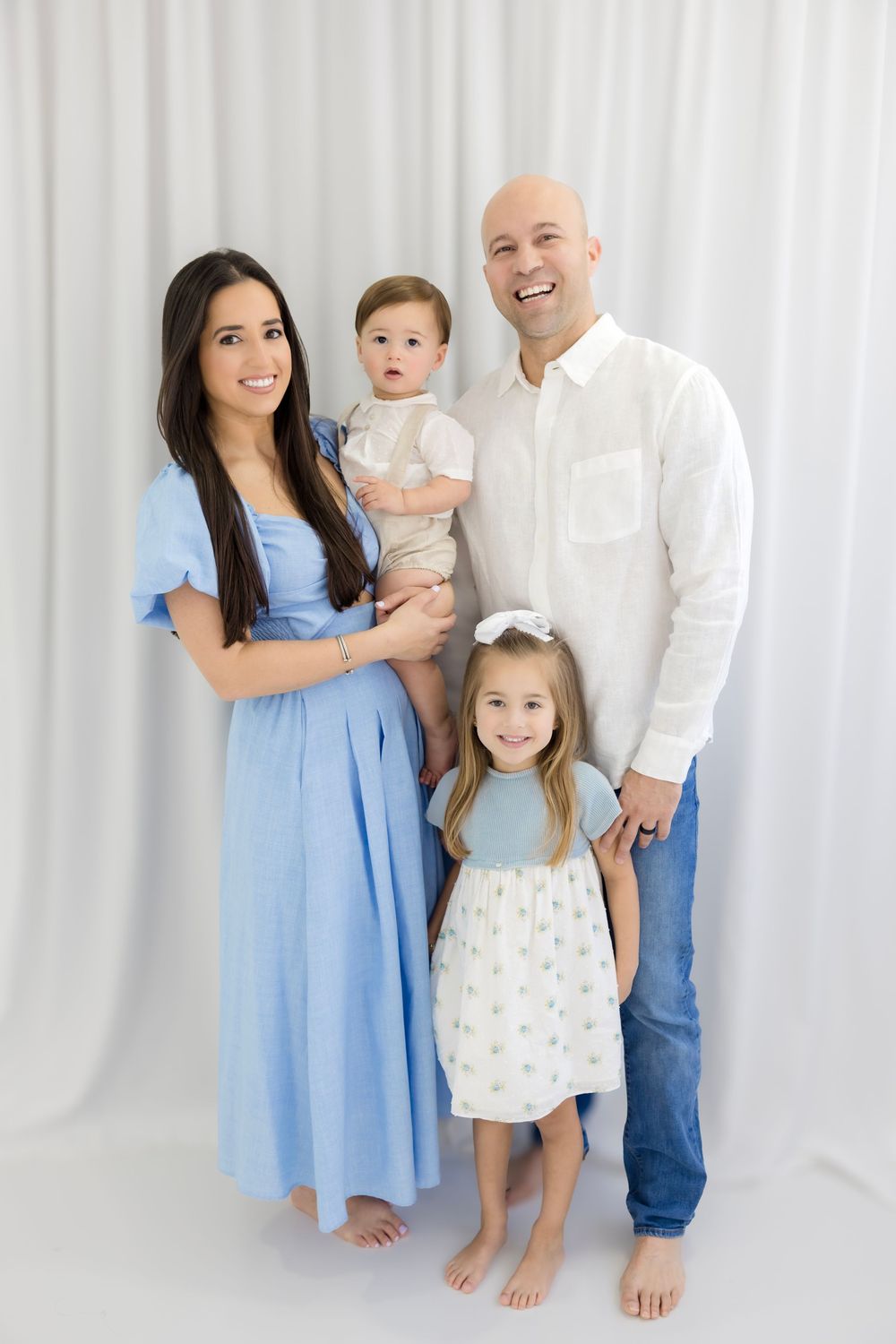 Happy family of four posing for a portrait, parents with toddler girl and baby boy, wearing coordinated blue and white outfits.
