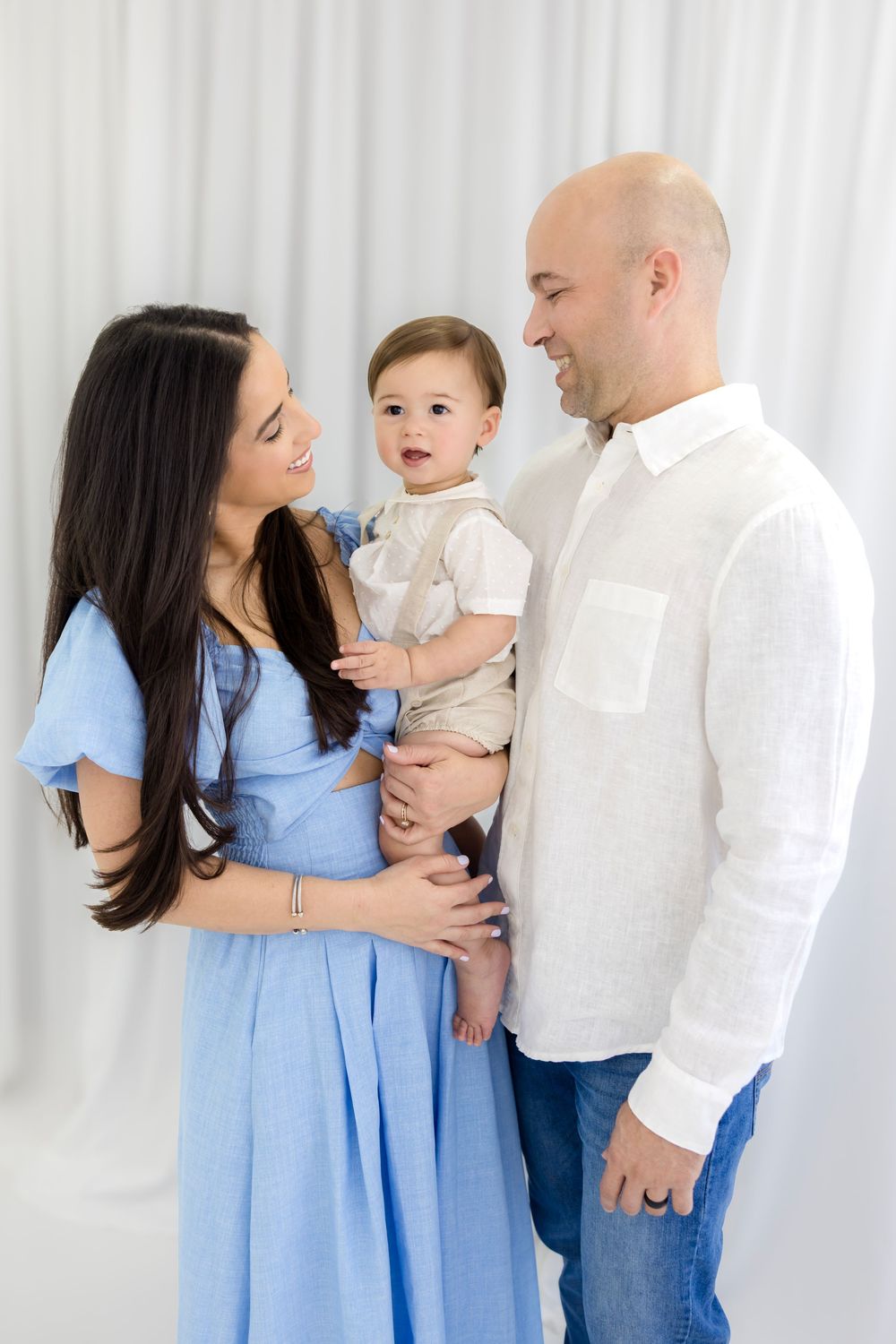 A happy young family with a bald father, dark-haired mother in blue dress, holding their baby boy in a bright white studio.