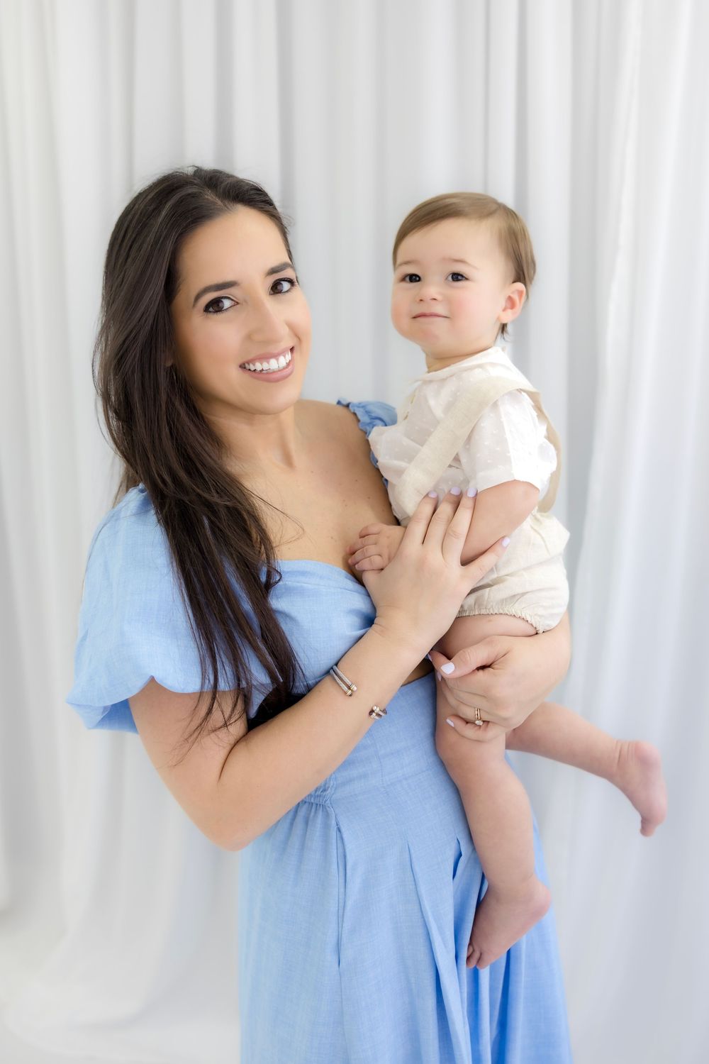 Smiling woman in blue dress holding a cute baby in white outfit against white curtain backdrop.