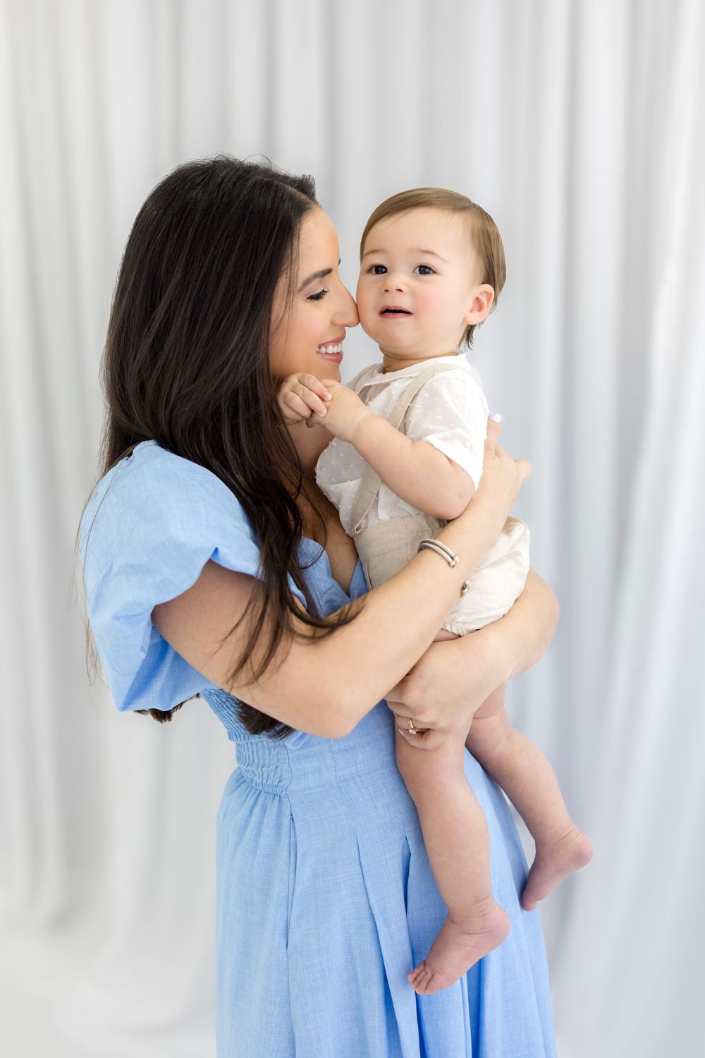 A smiling woman in a blue dress holds a happy baby in a white onesie against a light backdrop.